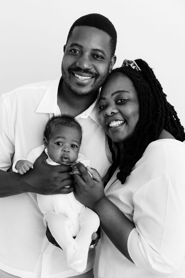 Black and white portrait of smiling parents holding their newborn baby, dressed in white outfits.
