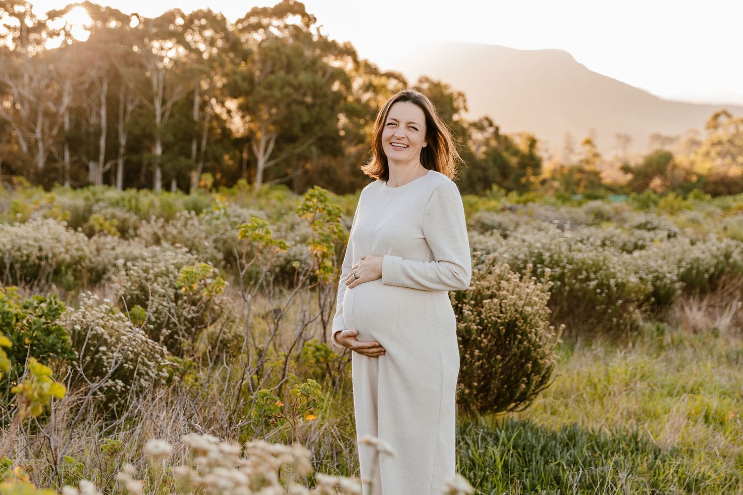 Smiling pregnant woman in white dress cradling baby bump outdoors at golden hour with mountains and greenery.