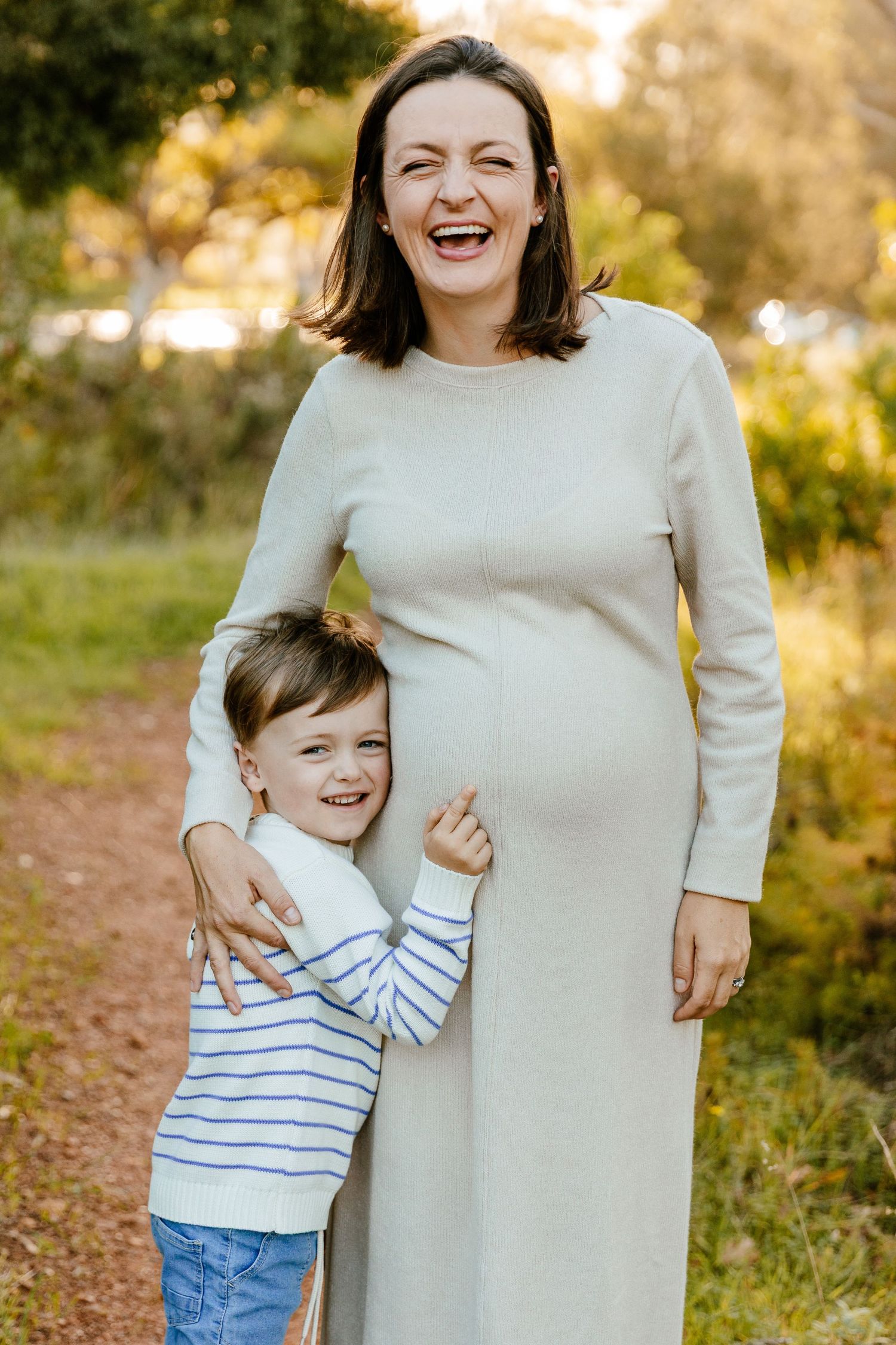 Pregnant woman laughing outdoors with young boy hugging her belly, both smiling in a sunlit park setting.