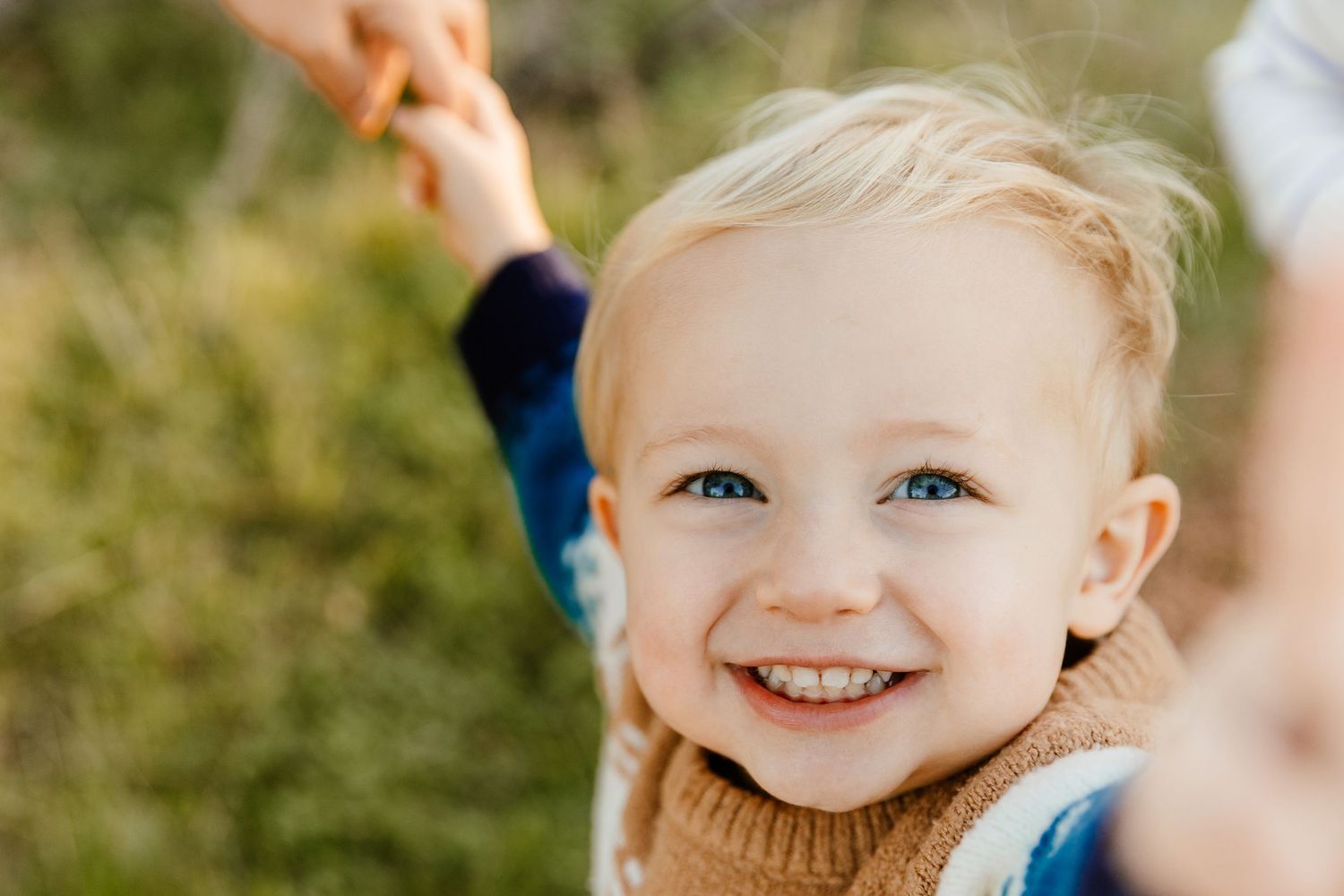 Smiling blonde toddler with blue eyes holding an adult's hand outdoors in a sunny natural setting.