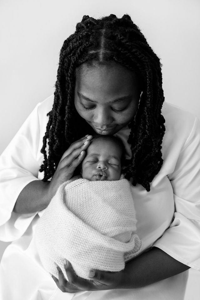 Black and white photo of a mother with braids tenderly kissing her swaddled newborn baby.