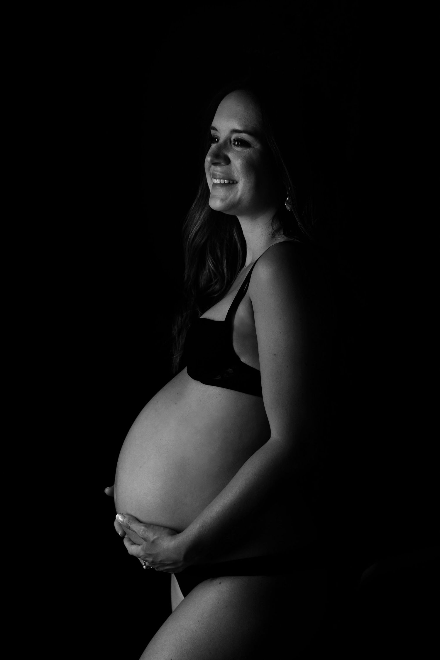 Black and white maternity photo of smiling pregnant woman cradling baby bump against dark background.
