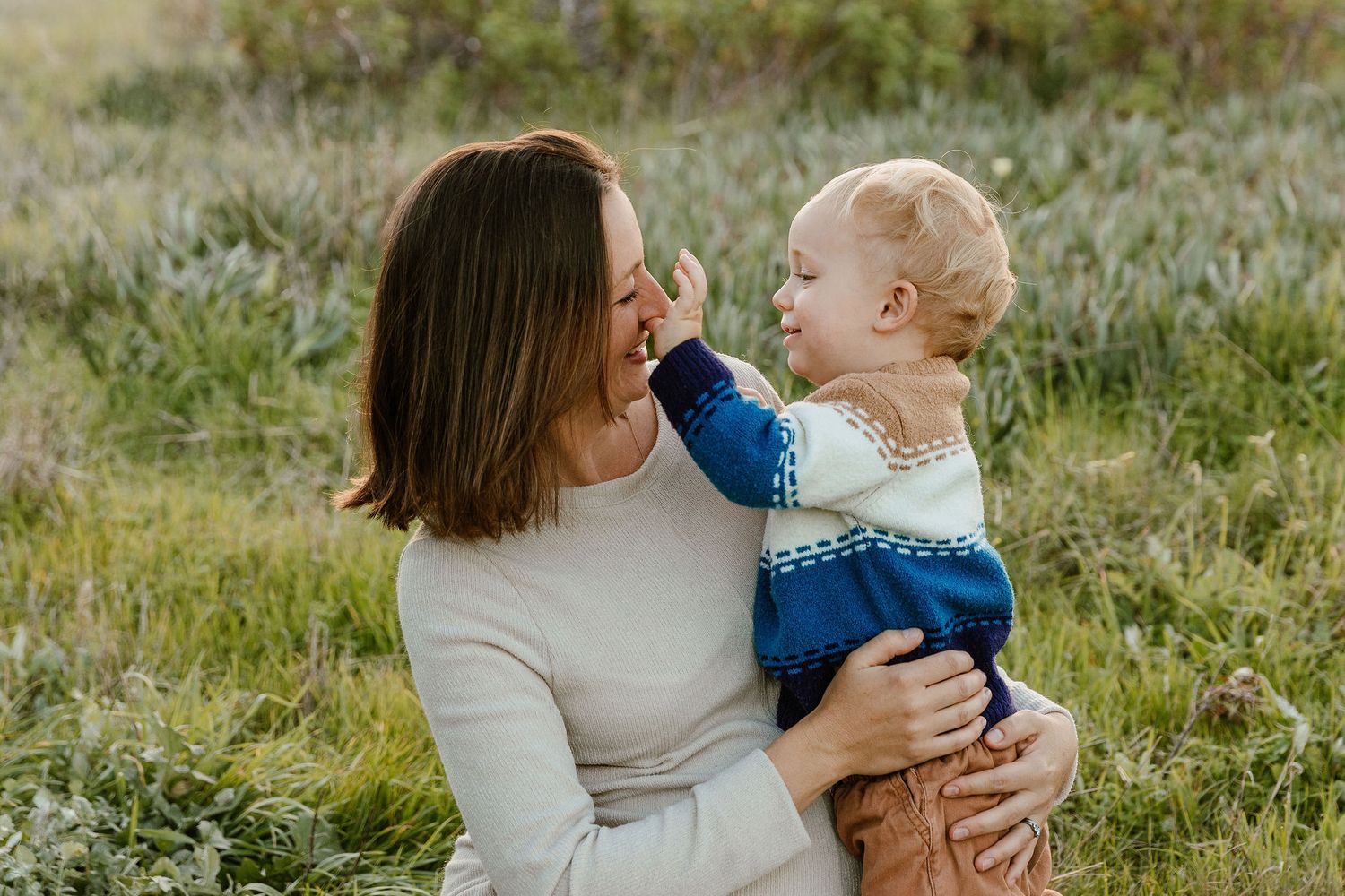 A smiling mother holds her blonde toddler who gently touches her face in a beautiful outdoor setting.