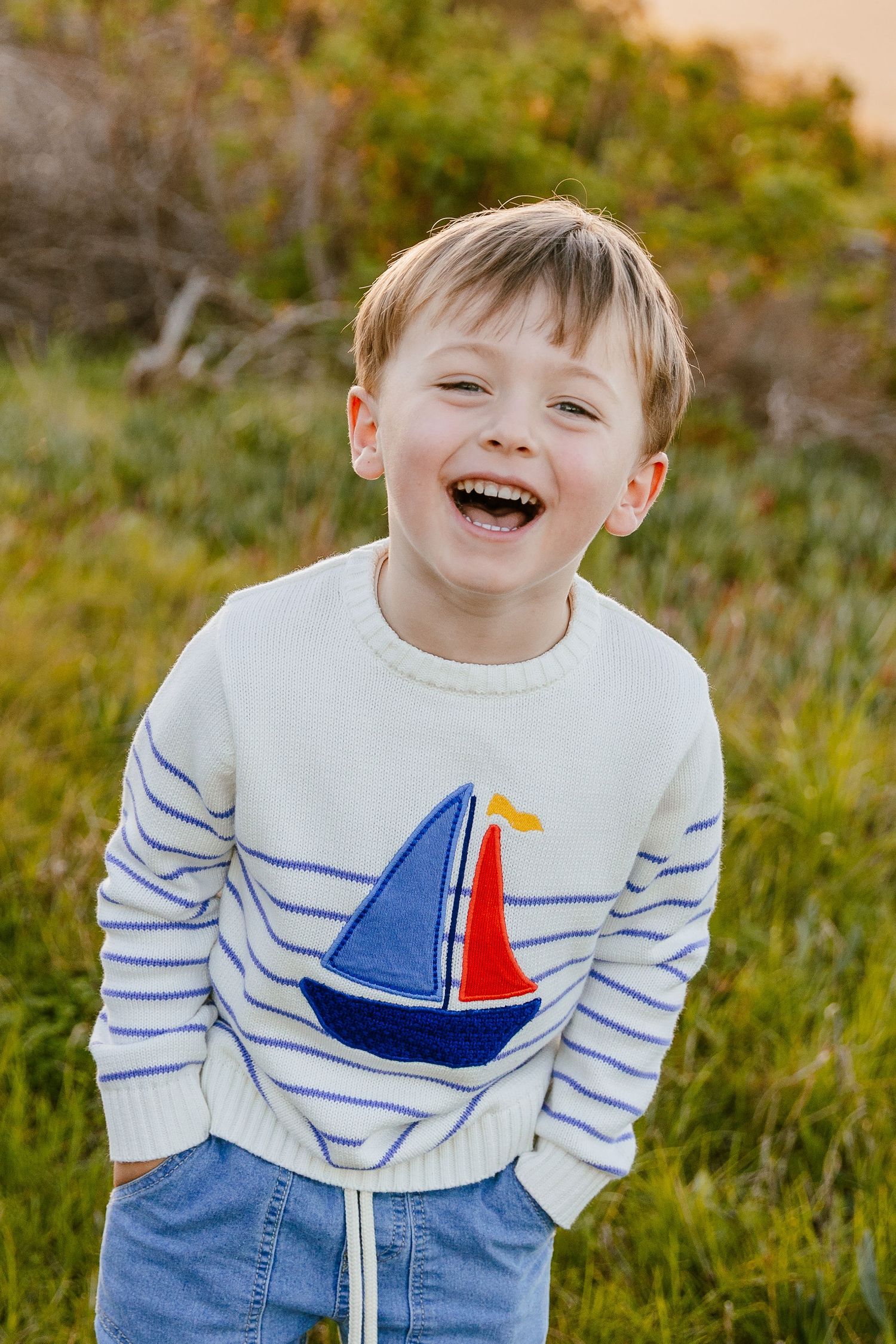 Laughing young boy in a blue striped sailboat sweater posing in a green field.