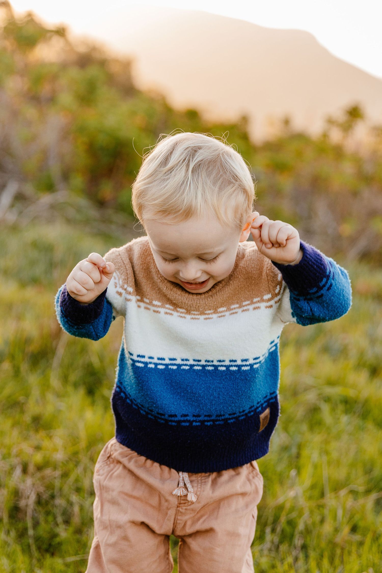 Toddler boy in colorful striped knit sweater laughing and playing outdoors in green field at golden hour.