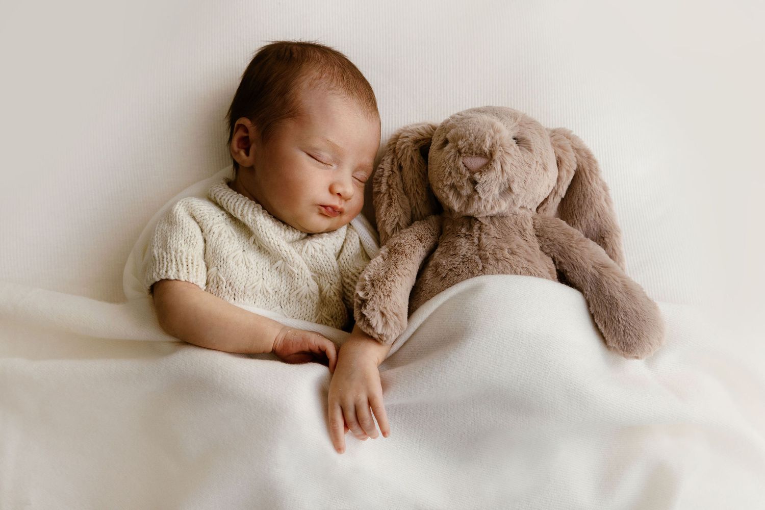 Sleeping newborn baby in a cream knit outfit cuddles with a soft brown bunny stuffed animal on white bedding.