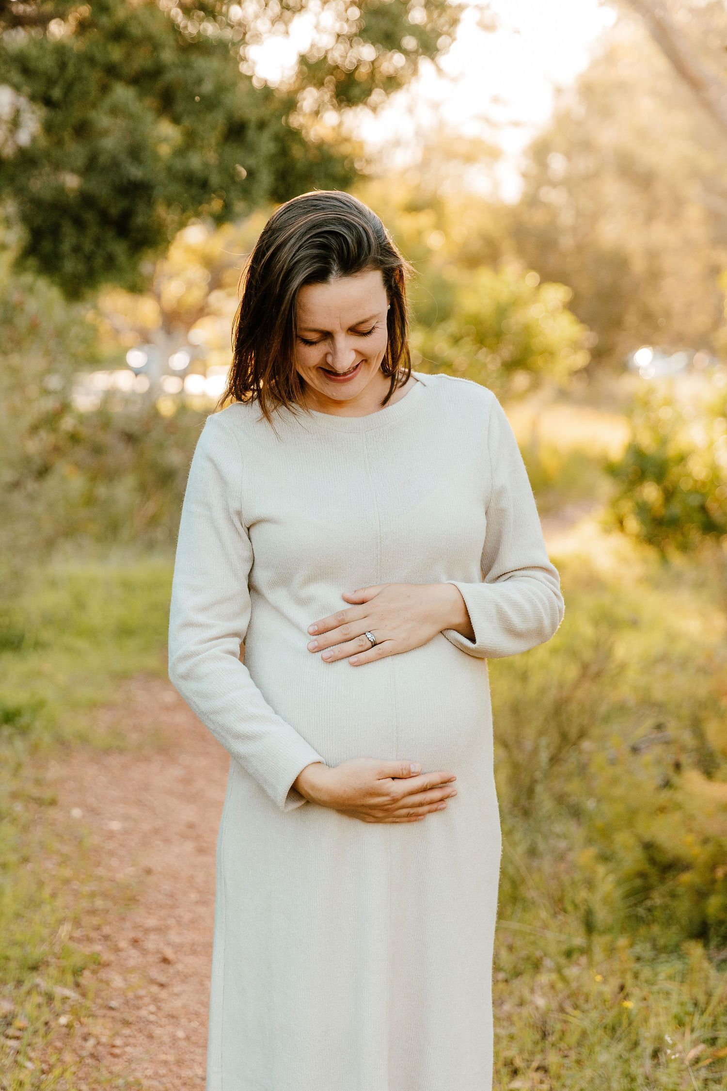 Smiling pregnant woman in white dress cradling baby bump outdoors in golden sunlight among lush greenery.