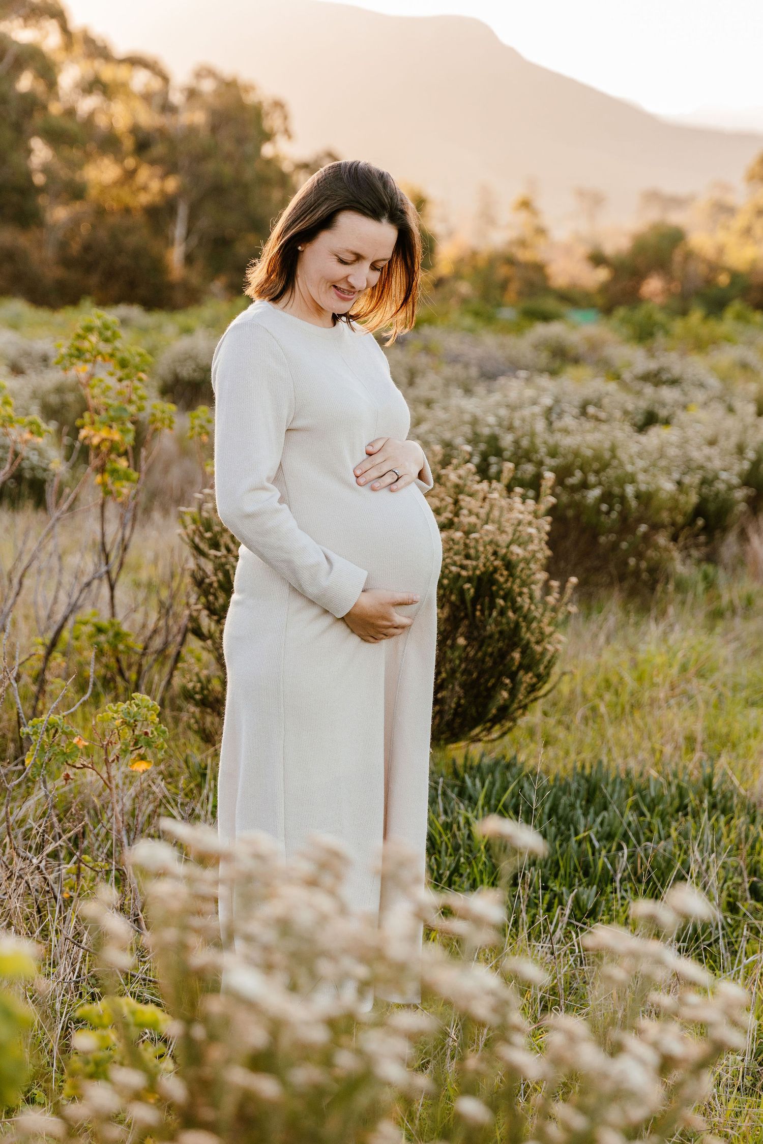 Smiling pregnant woman in white dress cradling her baby bump in a sunlit field with mountains in the background.