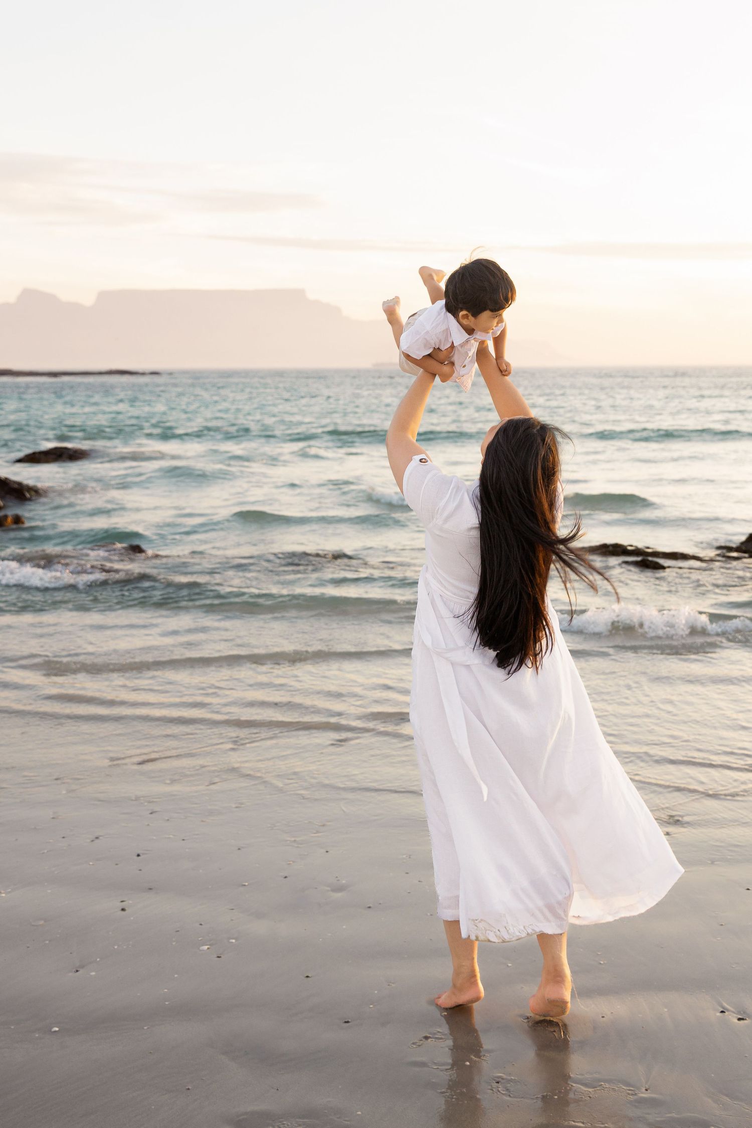 A mother in a white dress lifts her baby joyfully on a serene beach at sunset with ocean waves behind her.