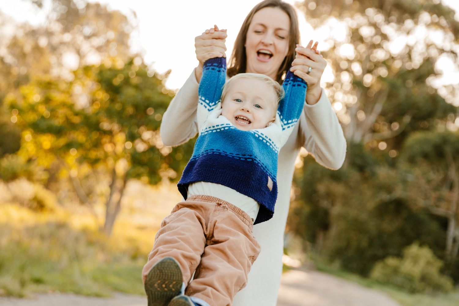 A joyful mother lifts her laughing toddler boy outdoors on a sunny path surrounded by green trees.