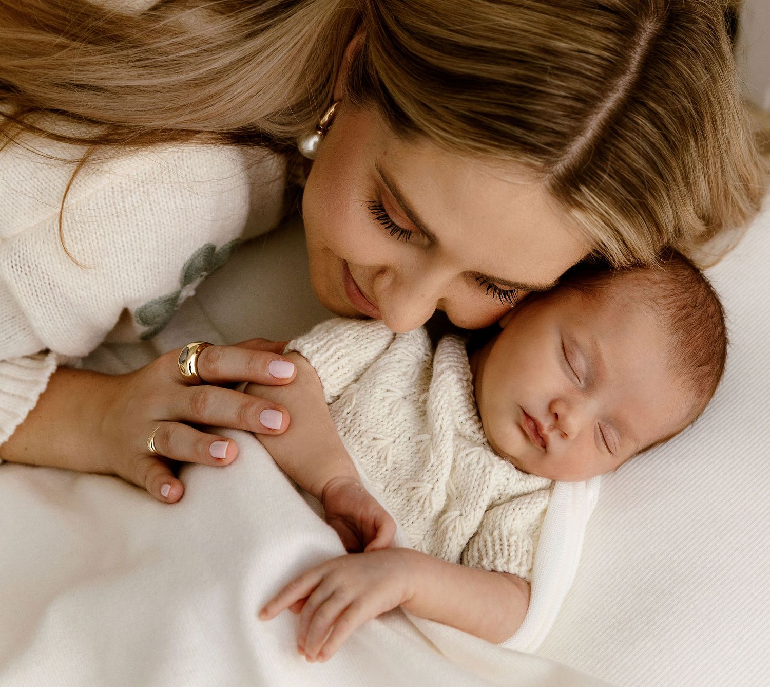 Mother tenderly kisses sleeping newborn baby wrapped in cream knit blanket.