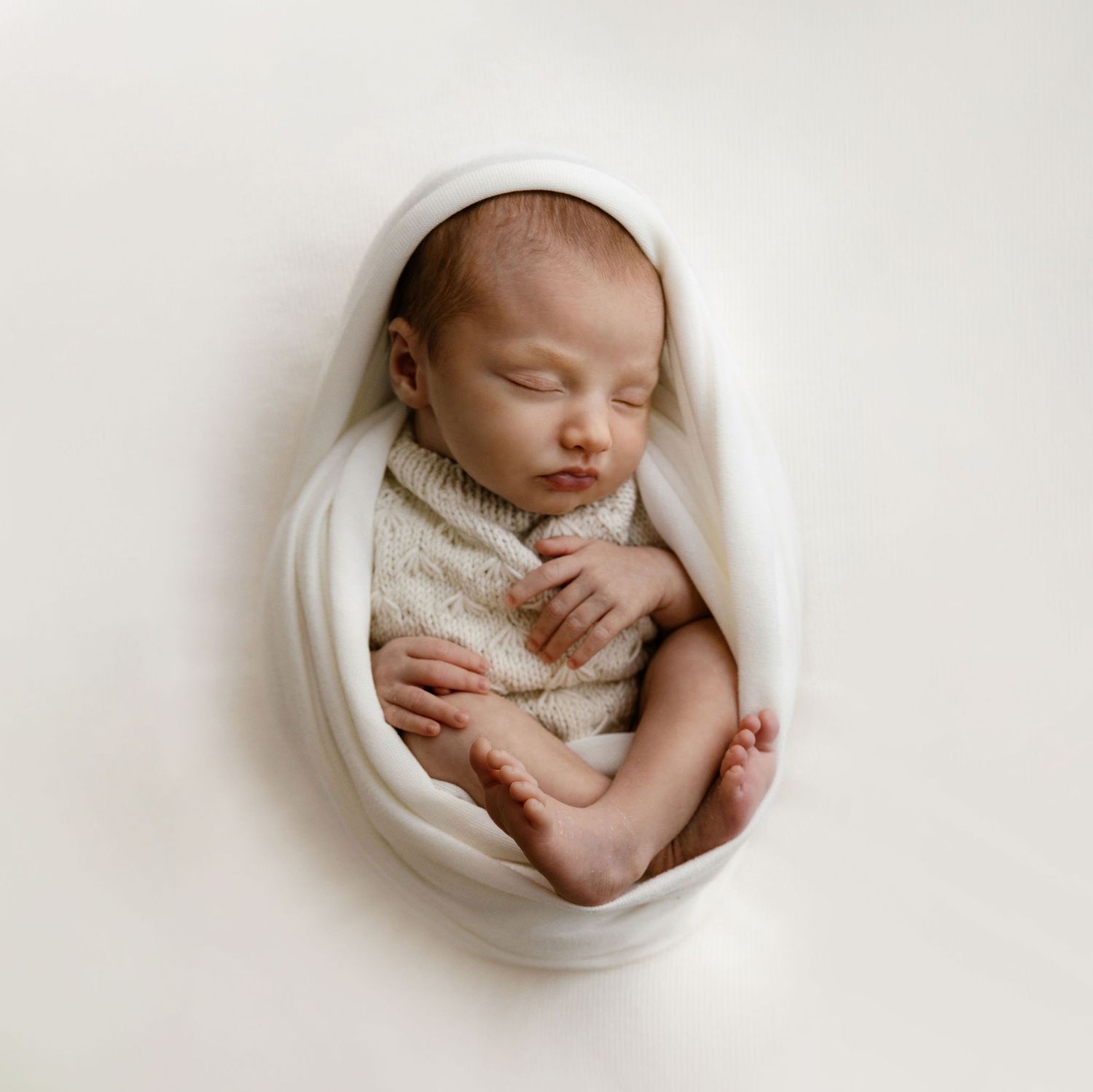 Newborn baby sleeping peacefully wrapped in soft white cloth in a cozy curled position.