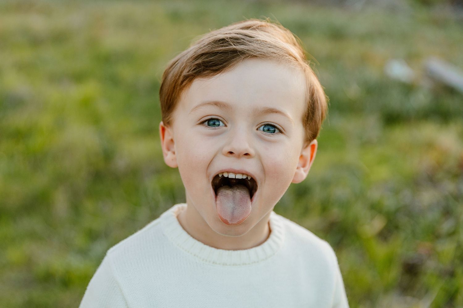 Playful young boy with blue eyes sticking his tongue out wearing a white sweater outdoors.