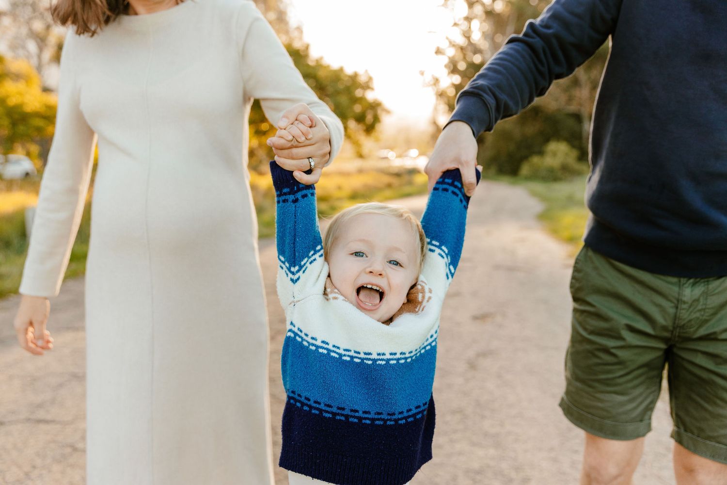 Happy toddler laughing with arms raised while parents hold hands on a warm sunset pathway.