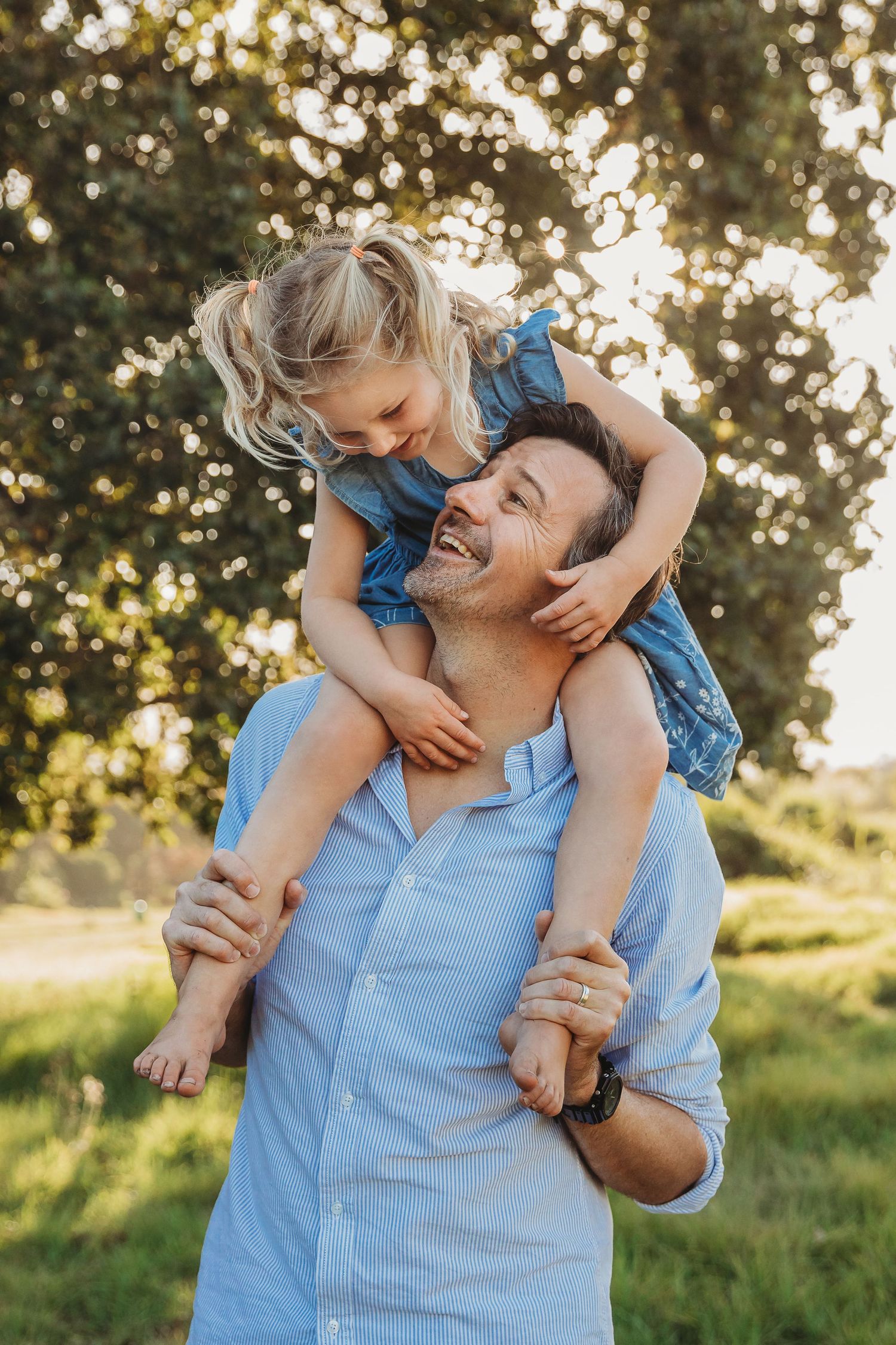 Smiling father carrying his young blonde daughter on his shoulders in a sunny green park.
