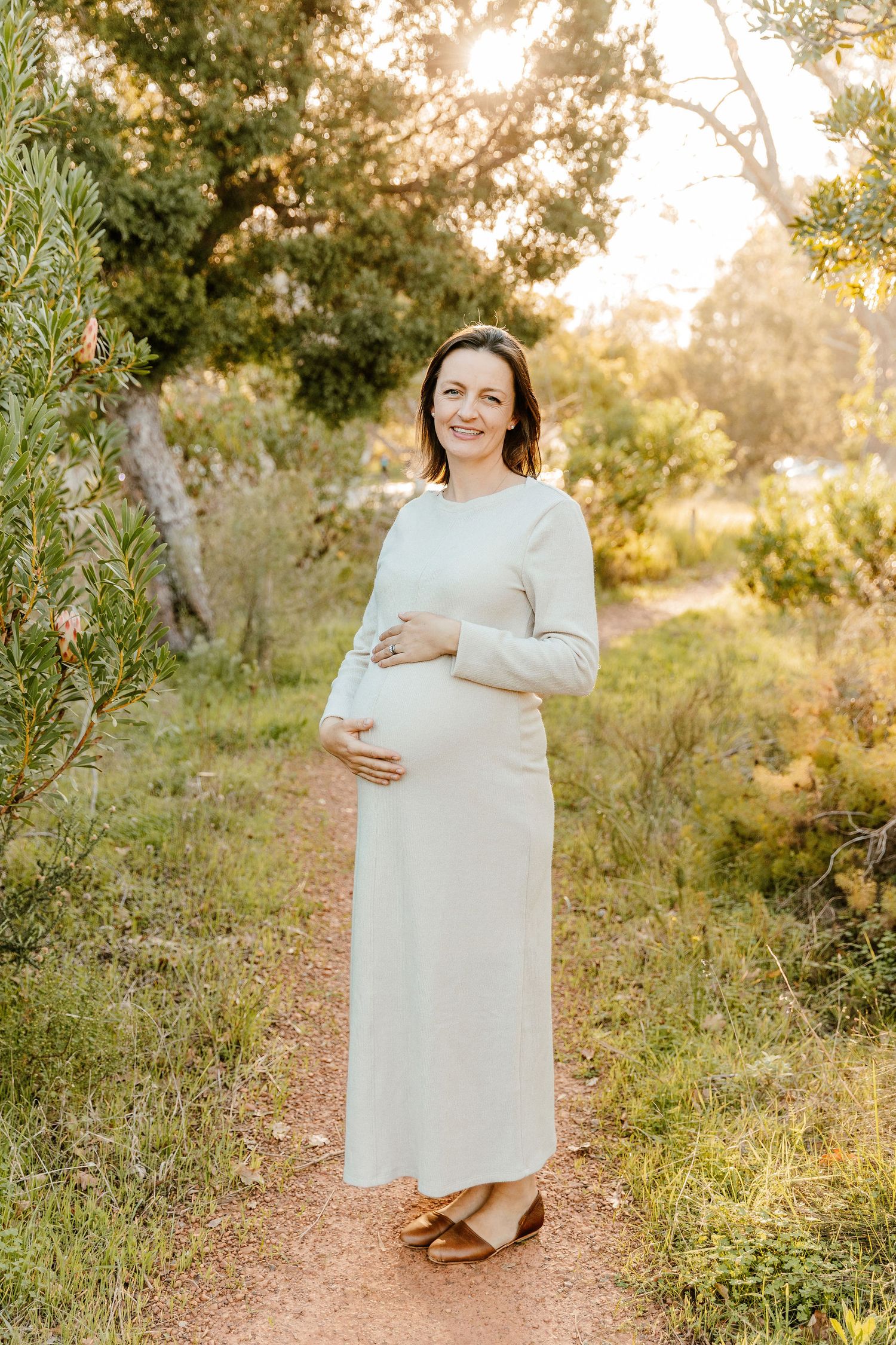 Smiling pregnant woman in a white maxi dress cradling her baby bump on a sunlit nature path.
