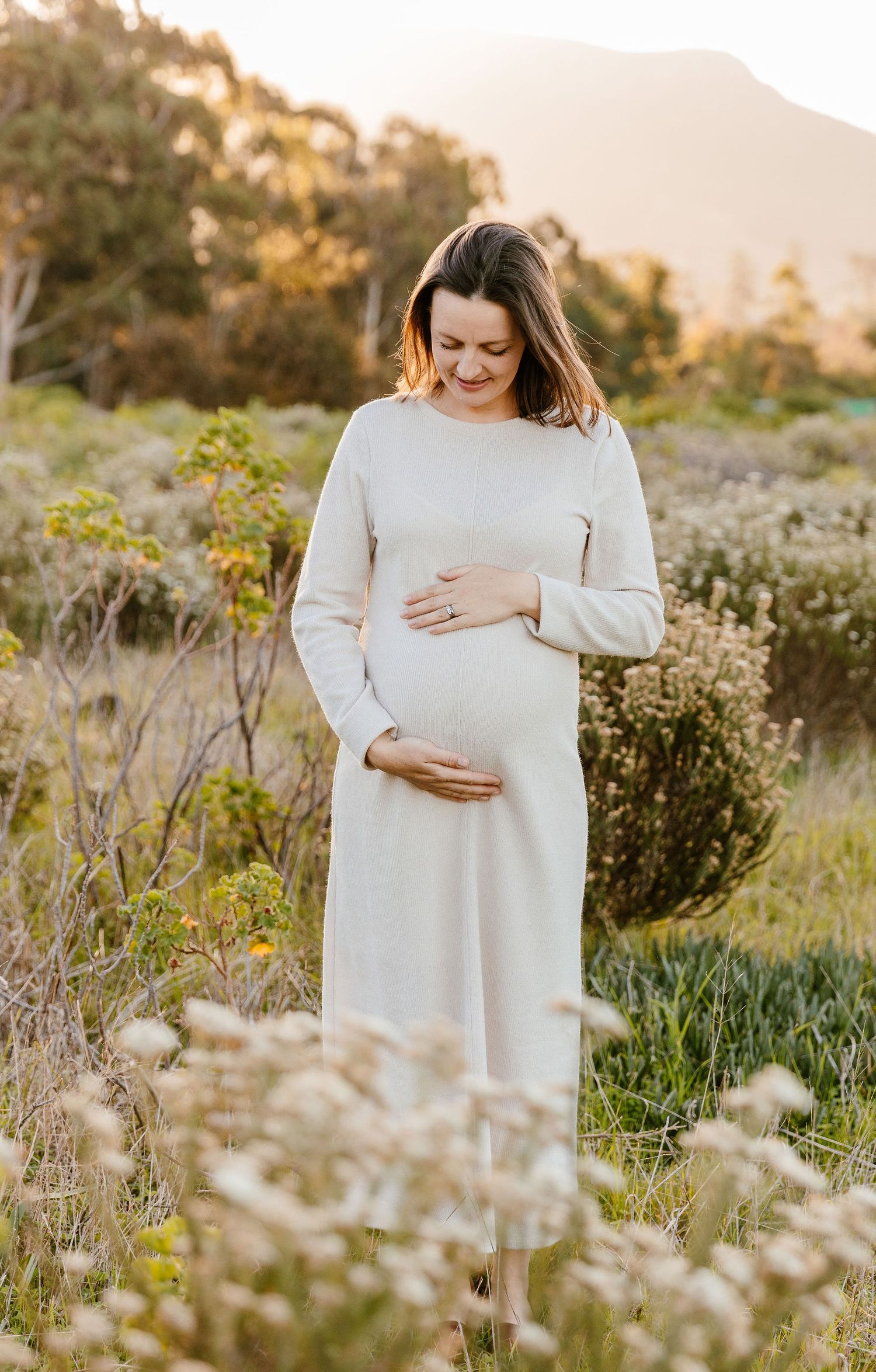 Pregnant woman in white dress cradling baby bump in sunlit field with mountains in background.