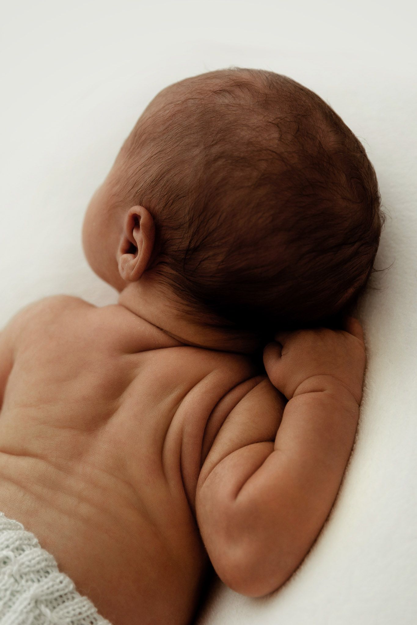 Newborn baby lying on back showing chubby arms and dark hair, peaceful infant portrait on white background.