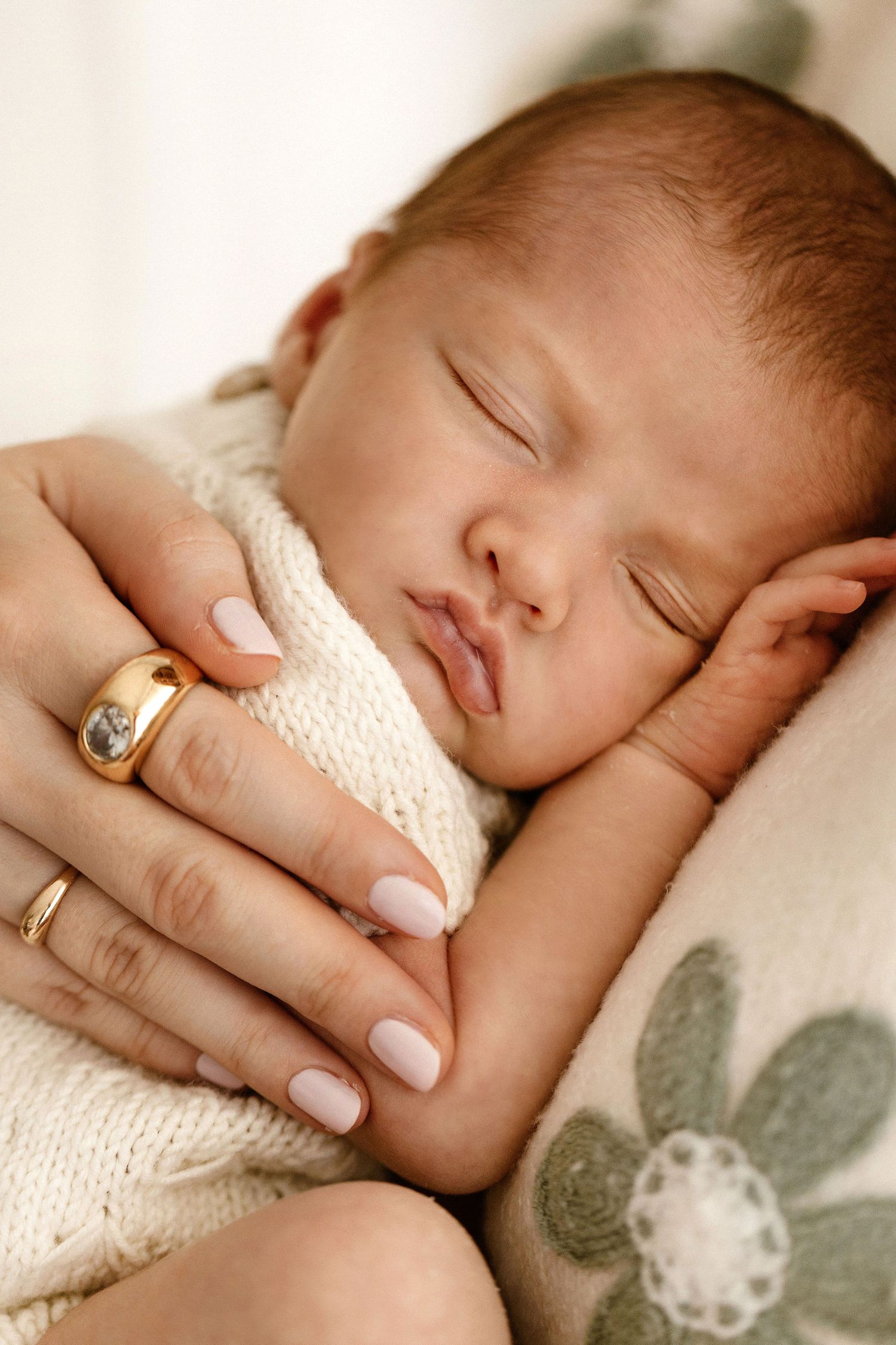 Sleeping newborn baby cradled in mother's hands, wearing a gold diamond ring, dressed in cream knit outfit.