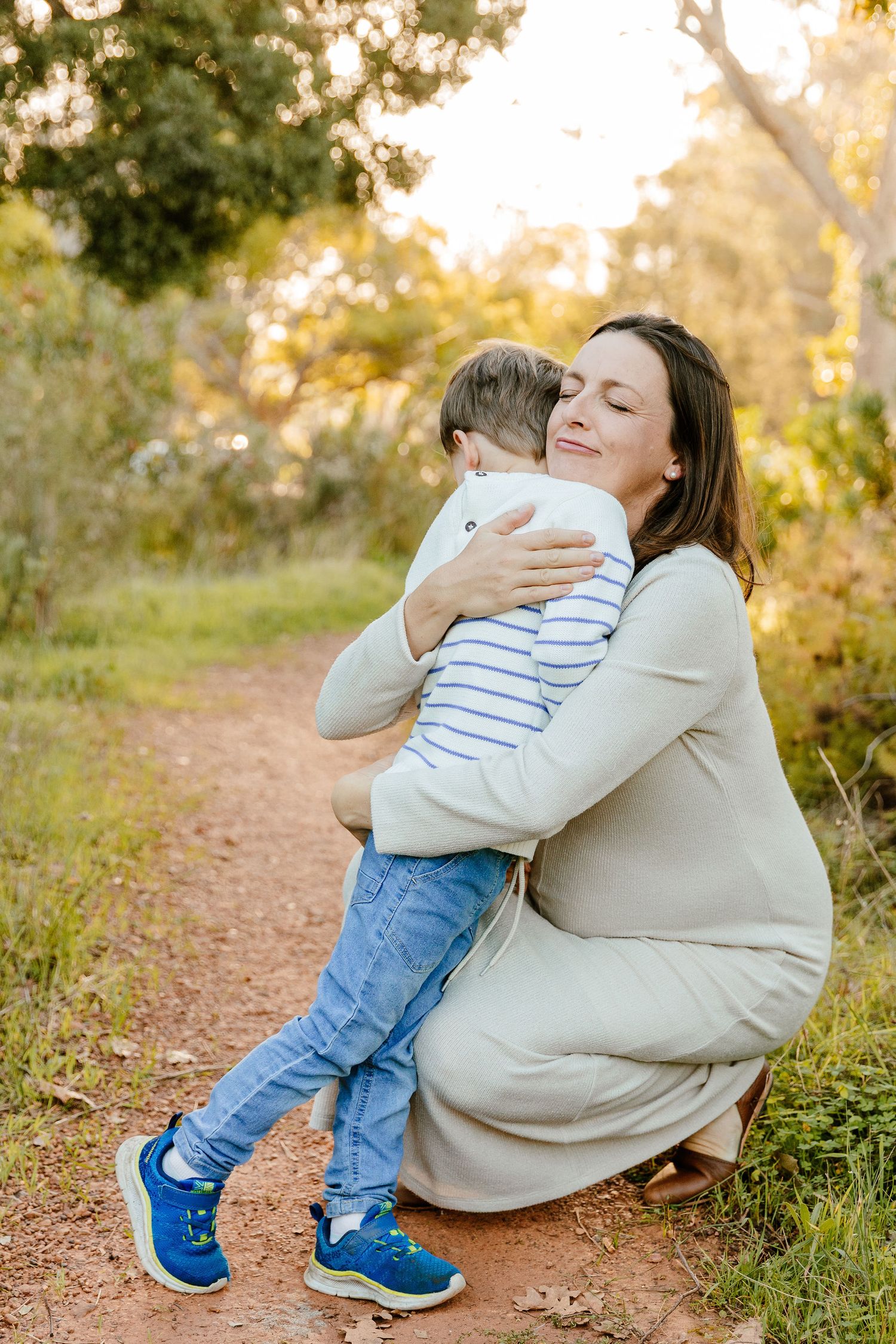 Pregnant woman in cream dress crouching to embrace young boy on a sunlit dirt path.
