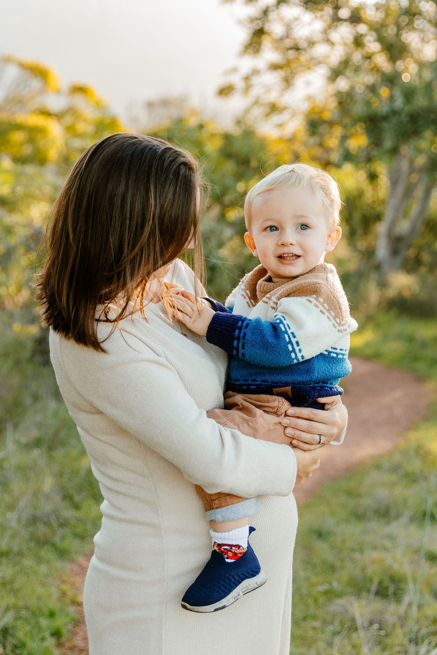 Mother in cream dress holding smiling blonde toddler on a nature trail at golden hour.
