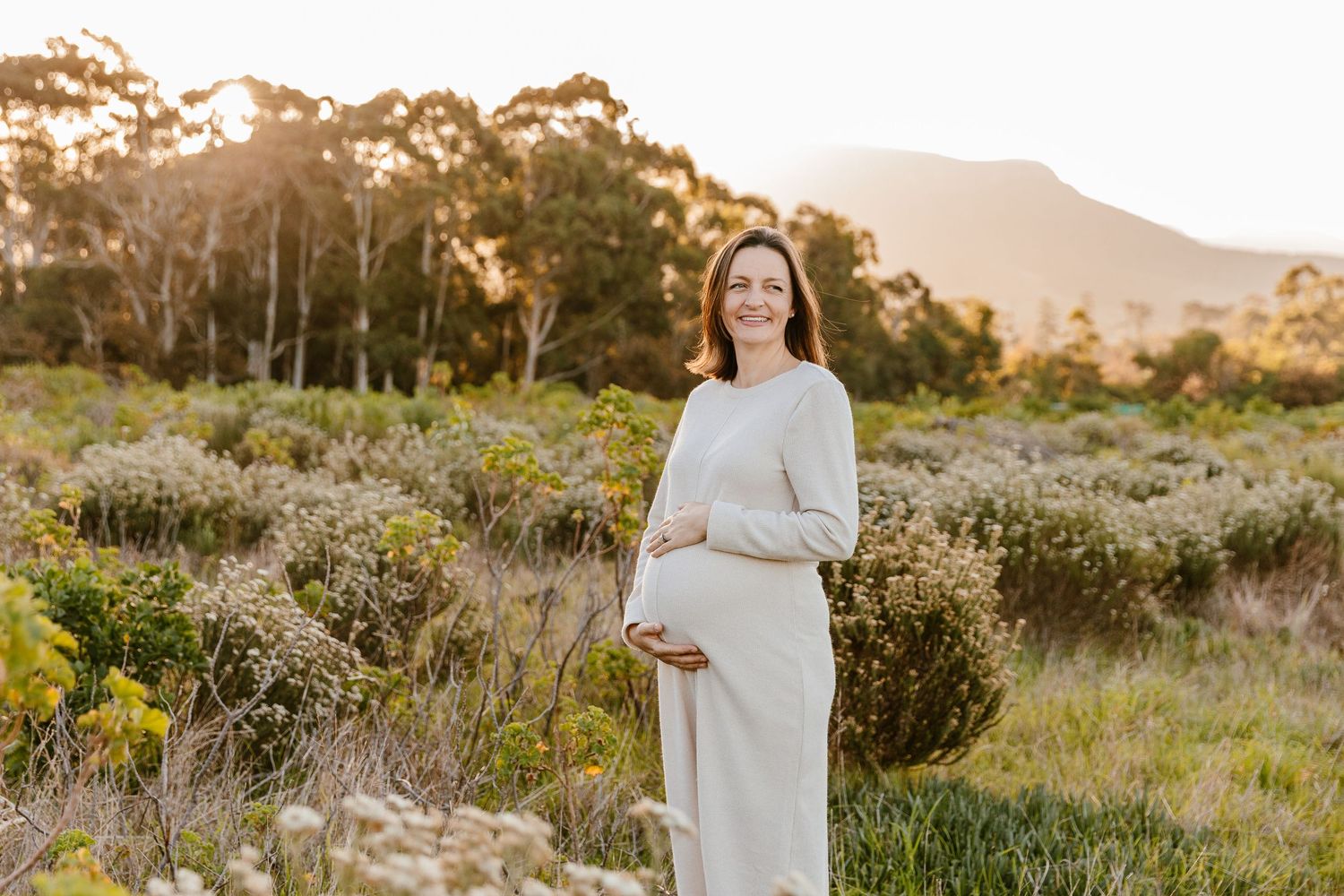 Smiling pregnant woman in white dress holding baby bump outdoors at golden hour with lush greenery and mountains behind.