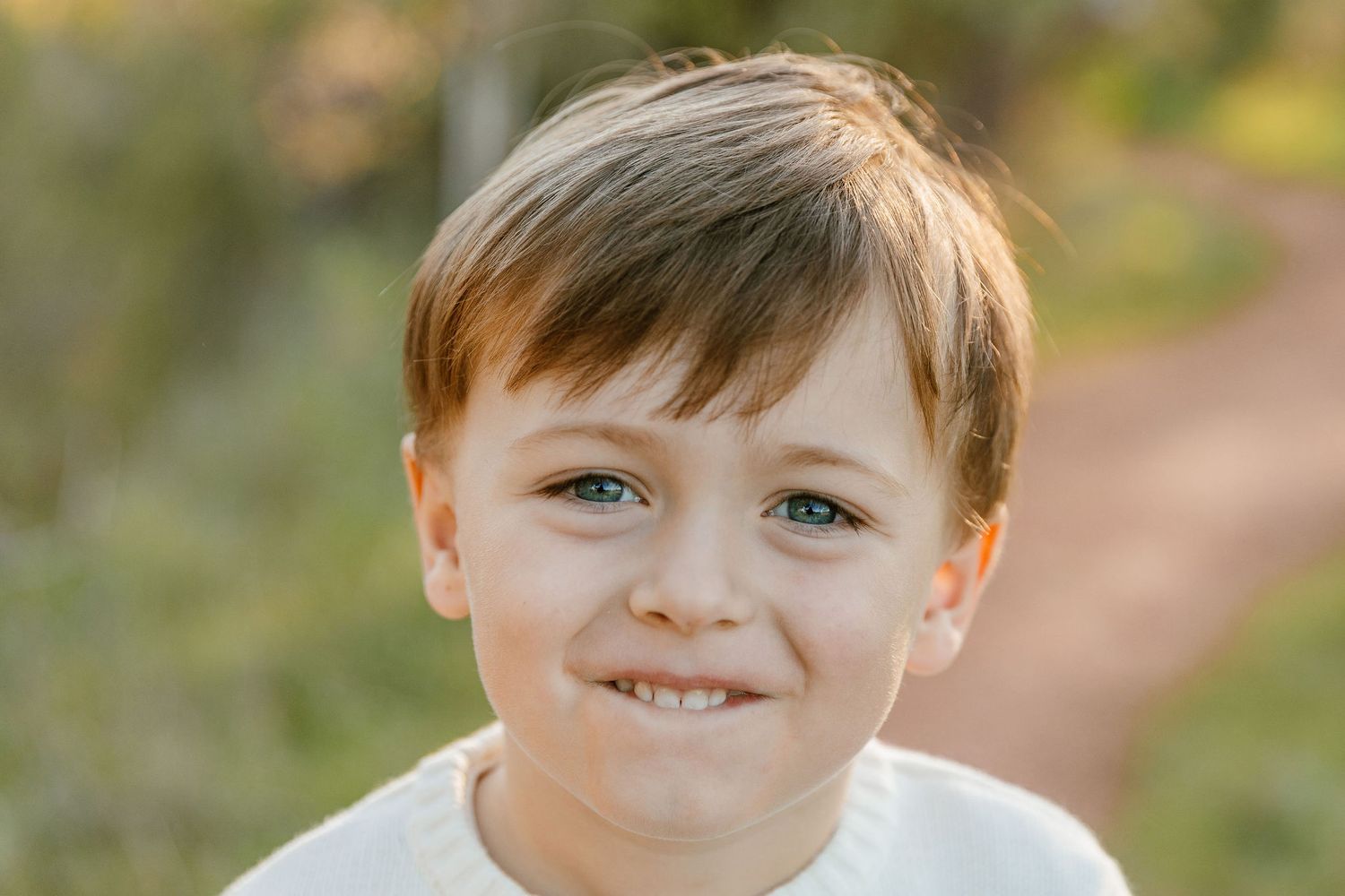 Smiling young boy with blue eyes and brown hair posing outdoors in white top.