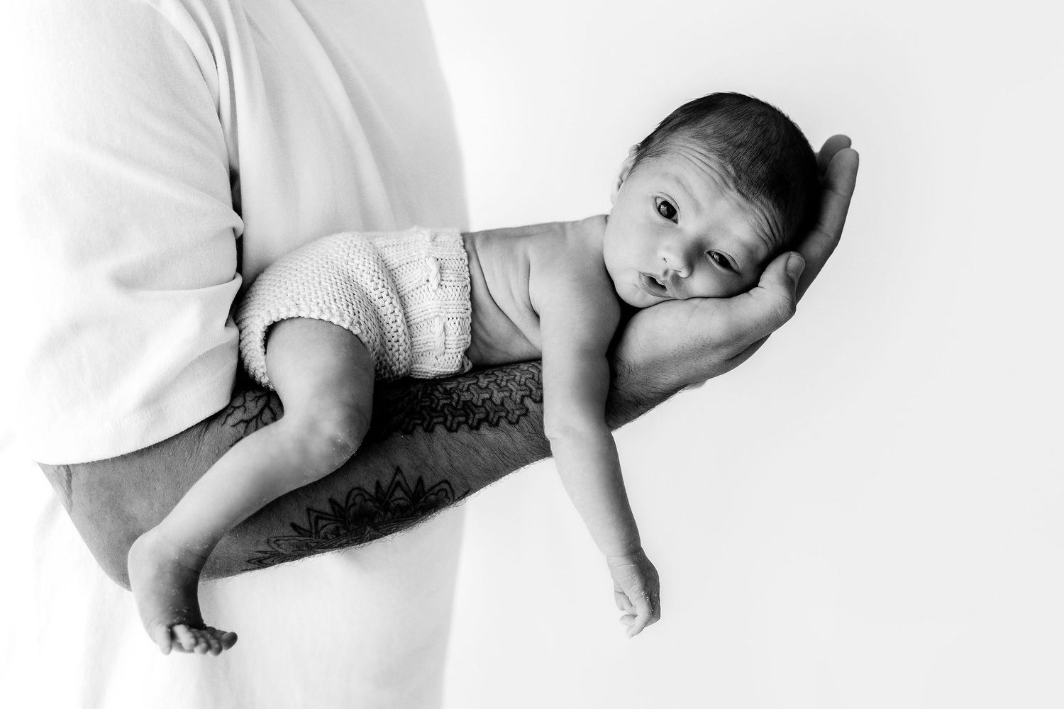 Black and white photo of newborn baby lying on tattooed father's forearm.