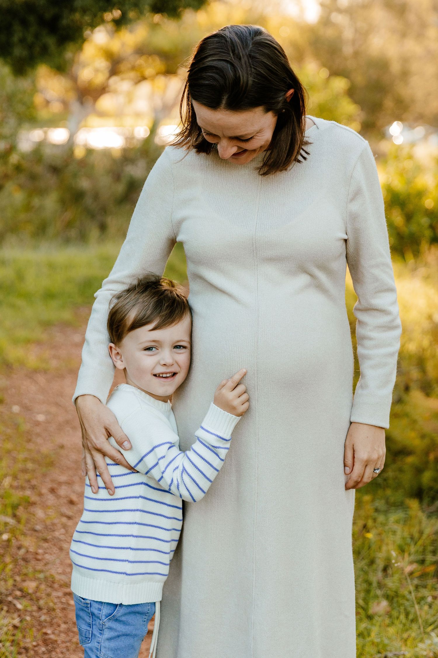 Pregnant woman in white dress smiling down at young child hugging her belly outdoors on a nature path.