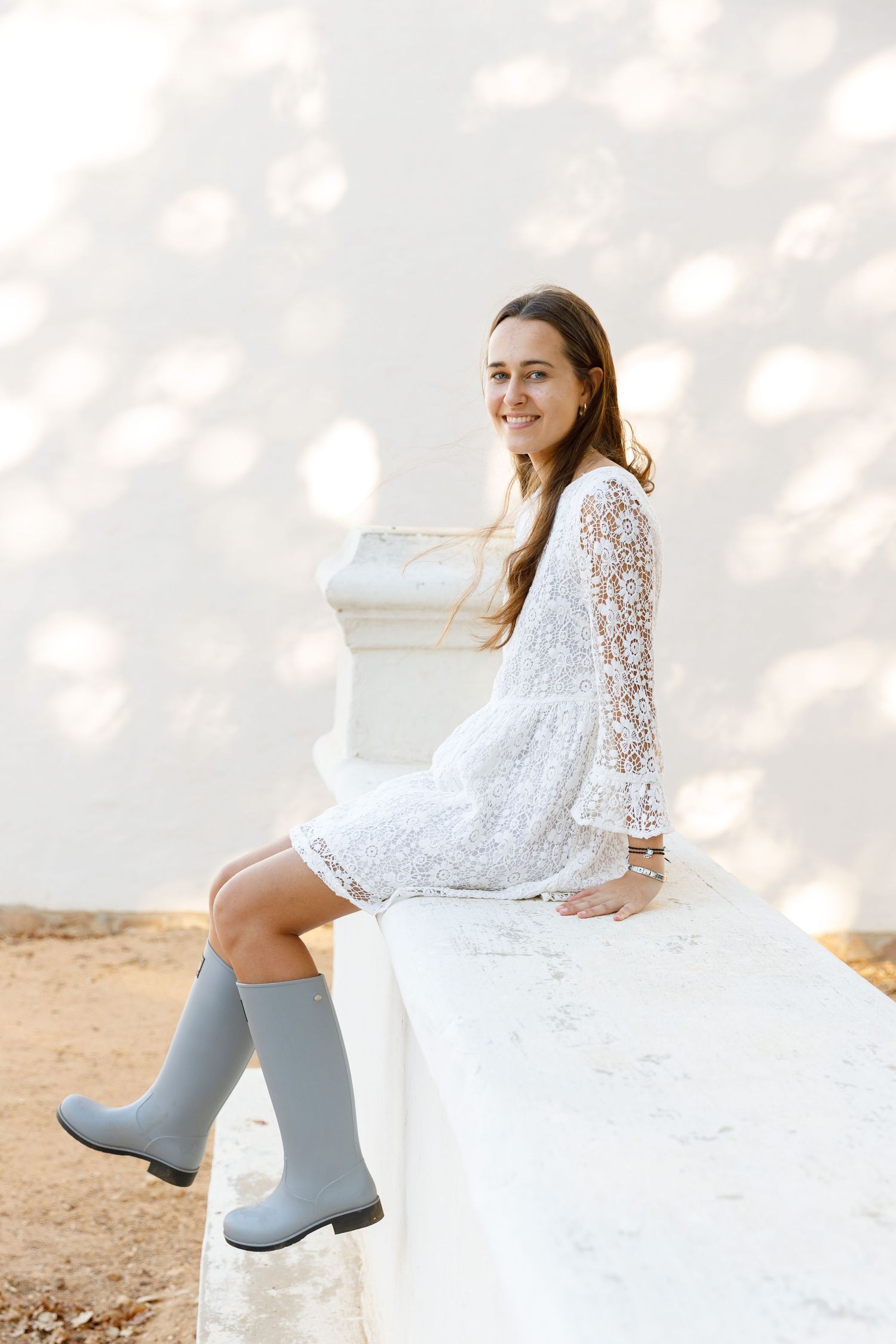 Young woman in white lace dress and gray rain boots sitting on white wall, smiling outdoors.