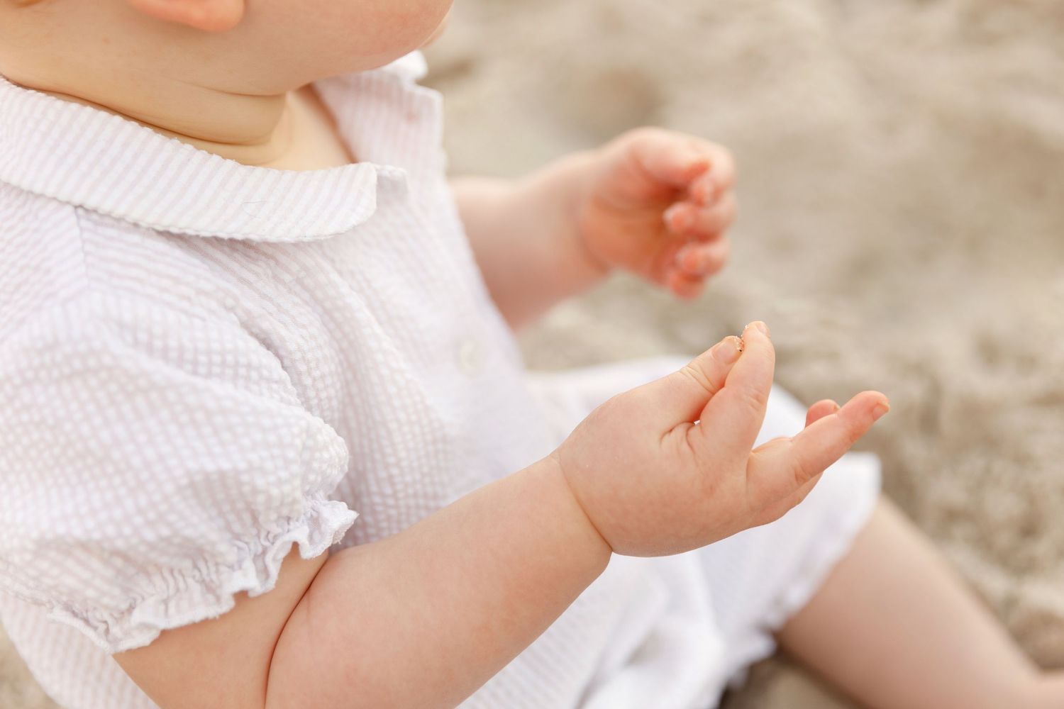 Close-up of a baby in white outfit sitting on sand, reaching out with tiny hands.