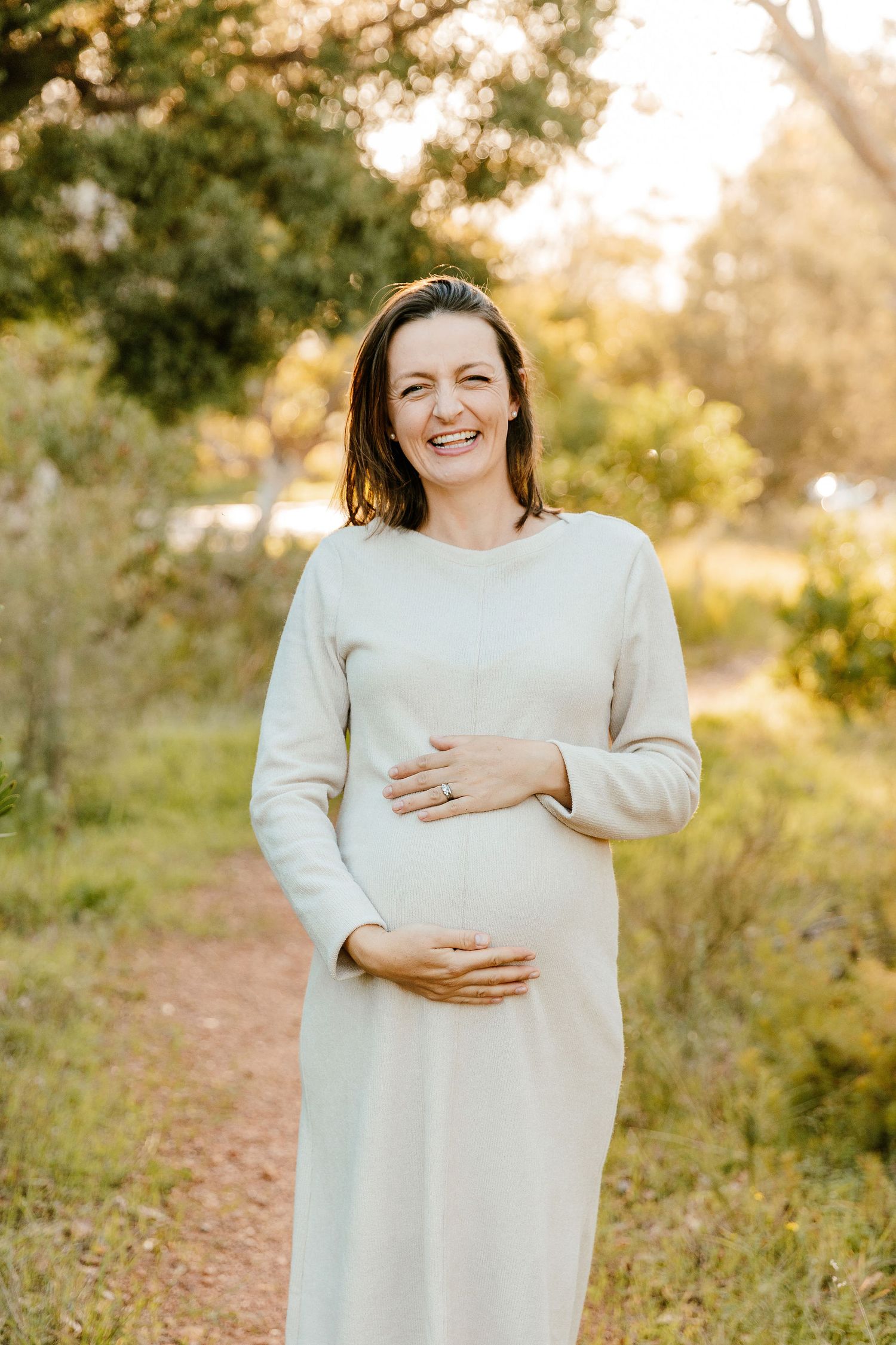 A smiling pregnant woman in a white dress cradles her baby bump on a sunny outdoor path.