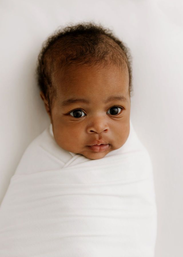 Newborn baby swaddled in white blanket looking up at camera with bright eyes and curly hair.