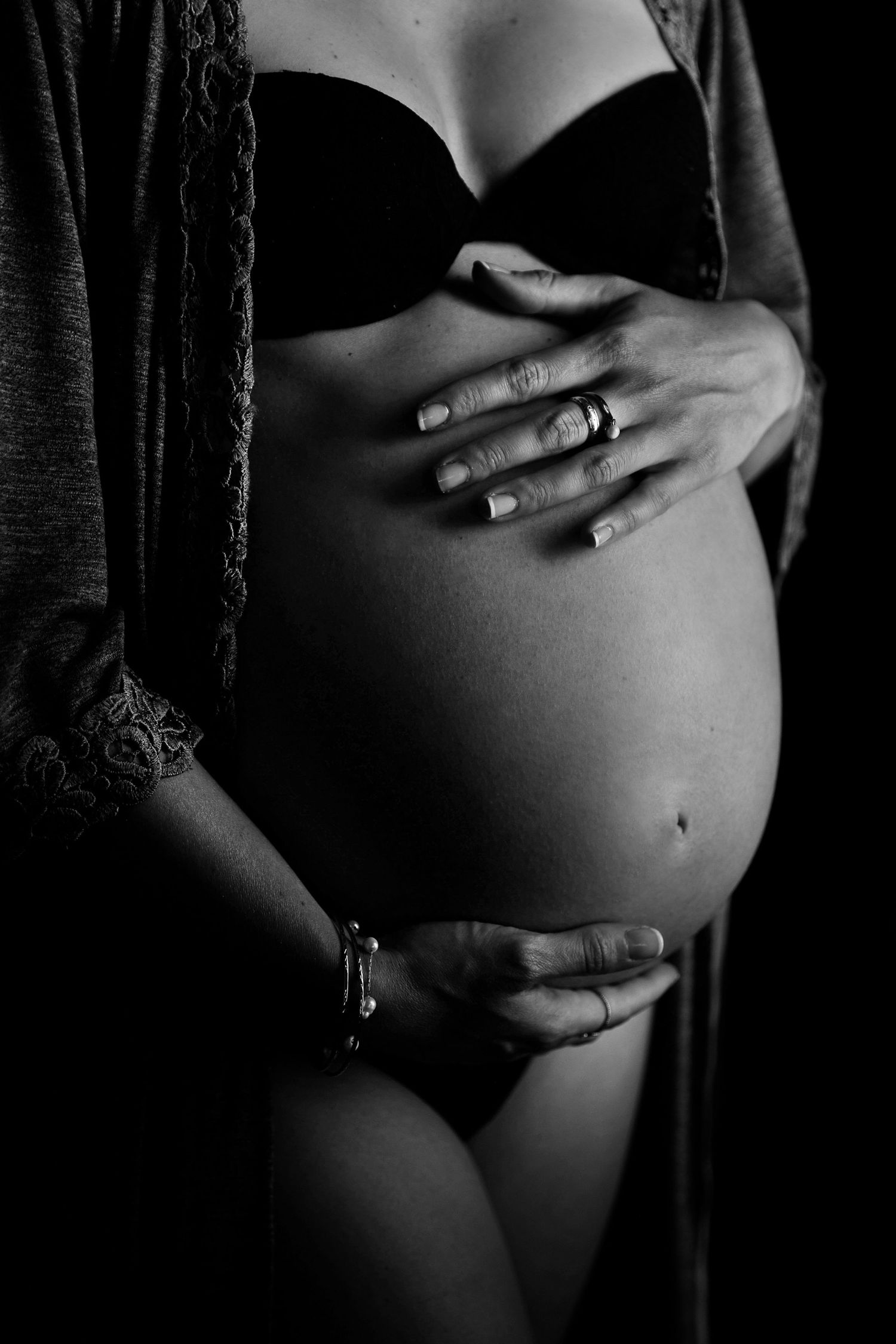 Black and white photo of pregnant woman cradling baby bump, wearing lingerie and open robe, showcasing maternity beauty.