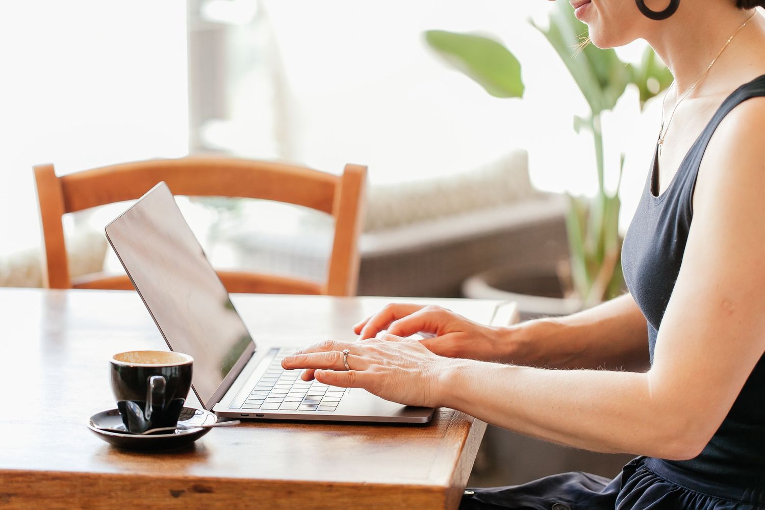 Woman typing on laptop at wooden table with coffee cup in bright cafe setting.