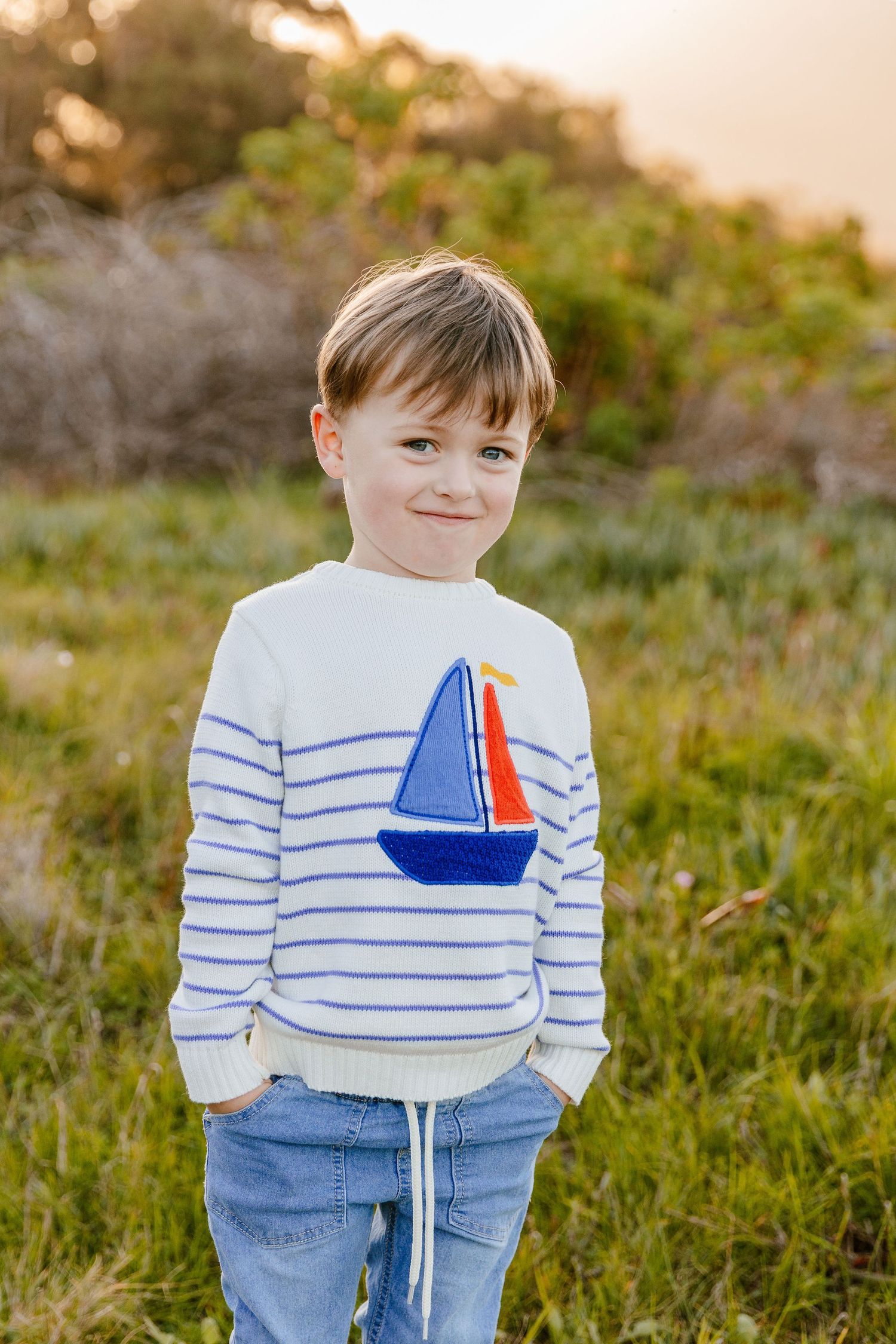Young boy in a striped sailboat sweater and jeans smiling in a sunny green field.