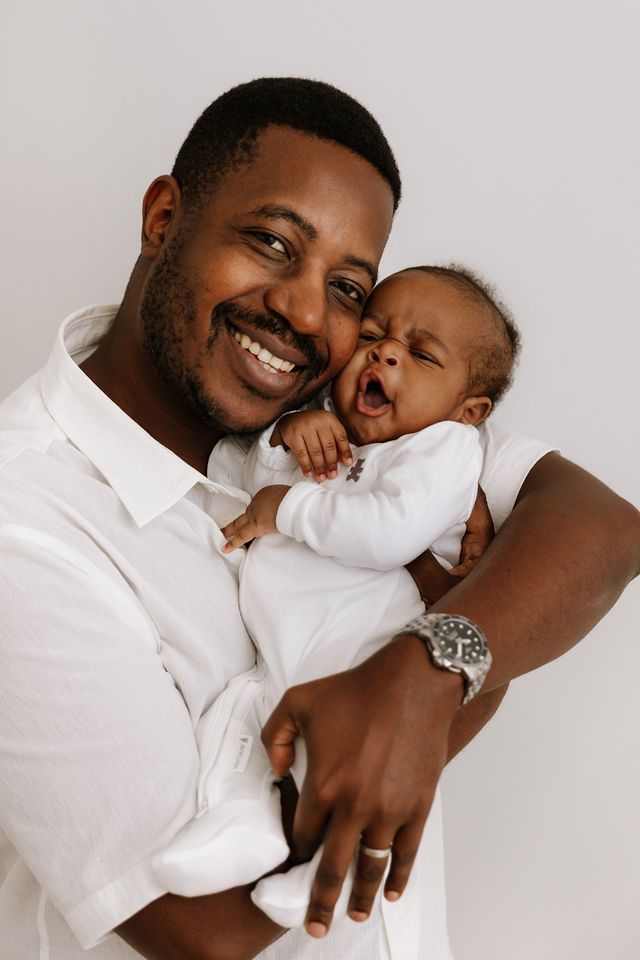 Smiling father in white shirt joyfully holds his yawning newborn baby against a light background.