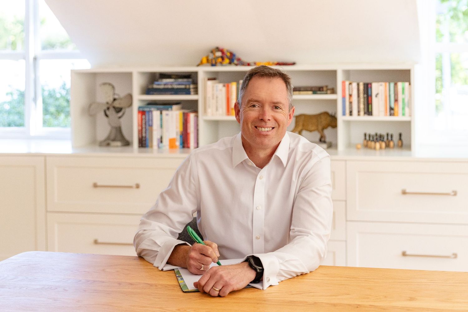Smiling businessman in white shirt sits at wooden desk writing notes in bright home office with bookshelves.