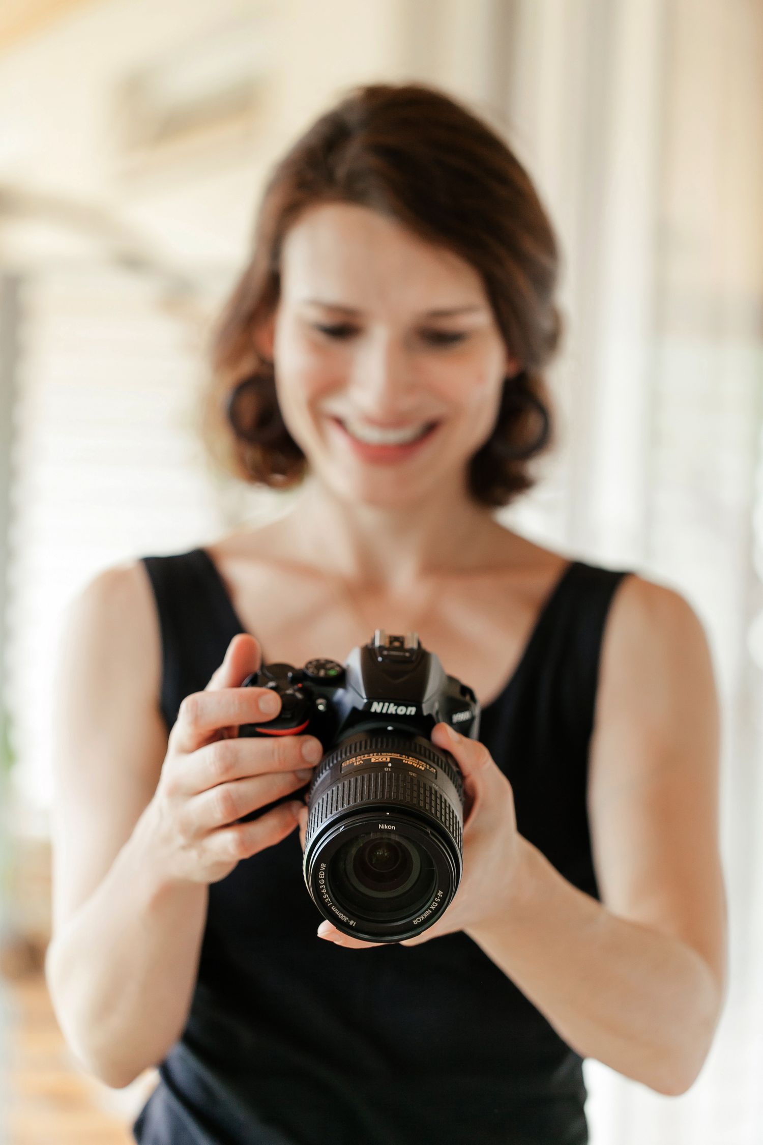 Smiling woman in black dress holding a Nikon DSLR camera, reviewing photos indoors with soft natural lighting.
