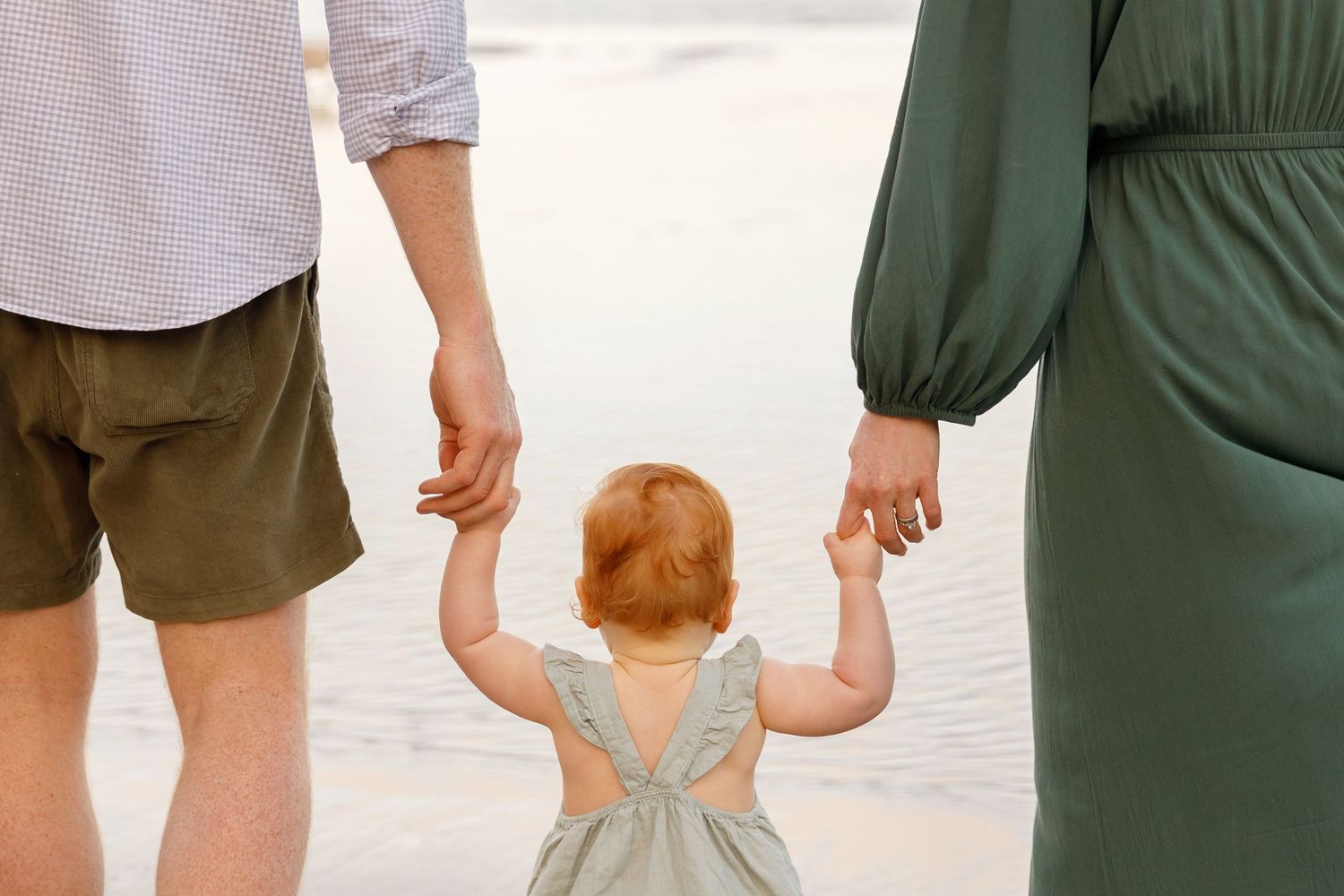 A toddler with red hair holds hands with parents on a beach, viewed from behind, wearing a gray dress.