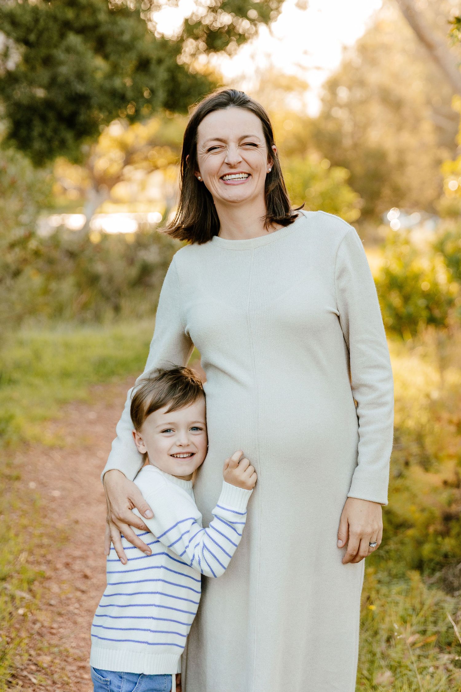 Pregnant woman in cream dress smiling outdoors with young boy hugging her belly on a sunny nature path.