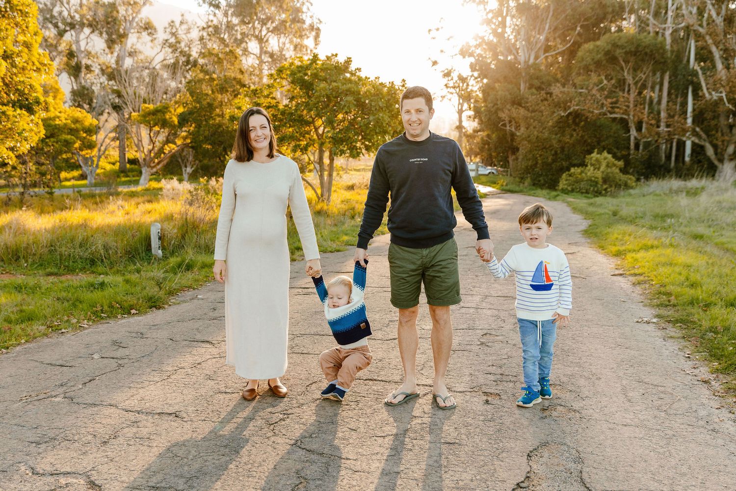 Pregnant mother, father, and two sons posing together on a sunny dirt road.