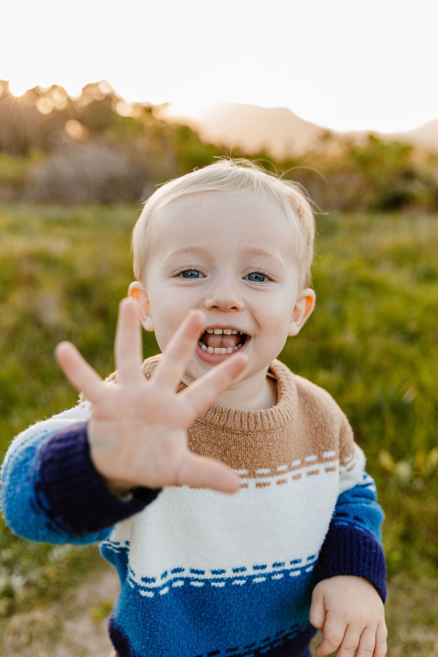 Excited blonde toddler in colorful knit sweater reaching toward camera in a sunny field.