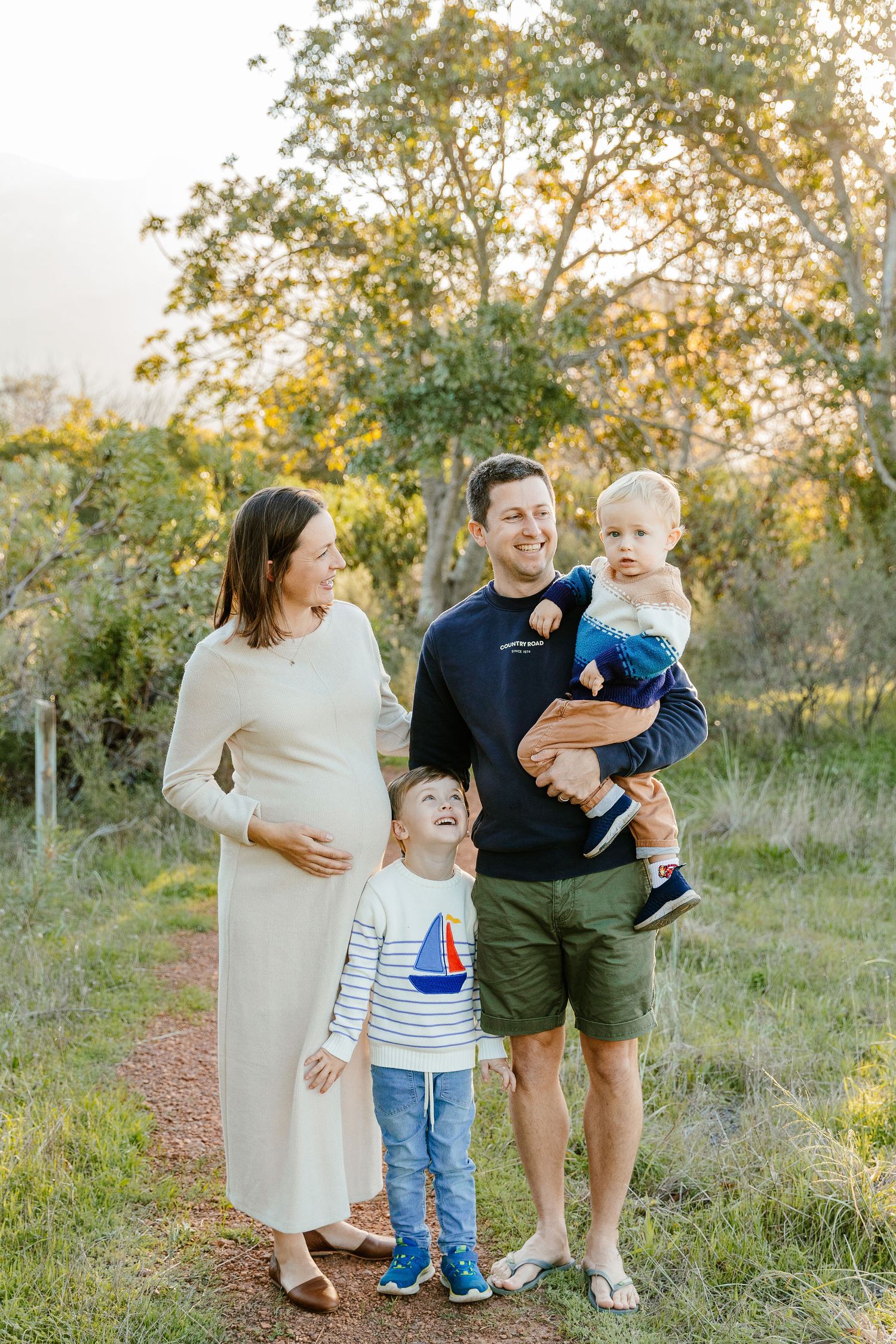Happy family of four outdoors at golden hour, pregnant mother in cream dress with father holding toddler and young son.