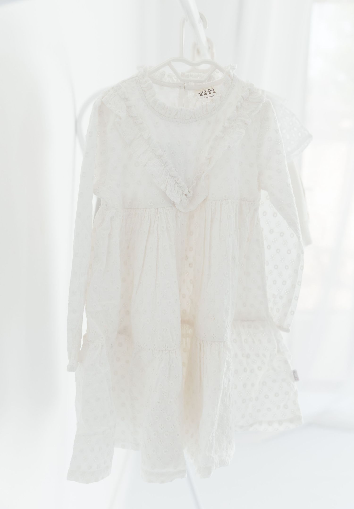 White lace tiered mini dress with long sleeves and ruffle details, hanging on a white hanger.