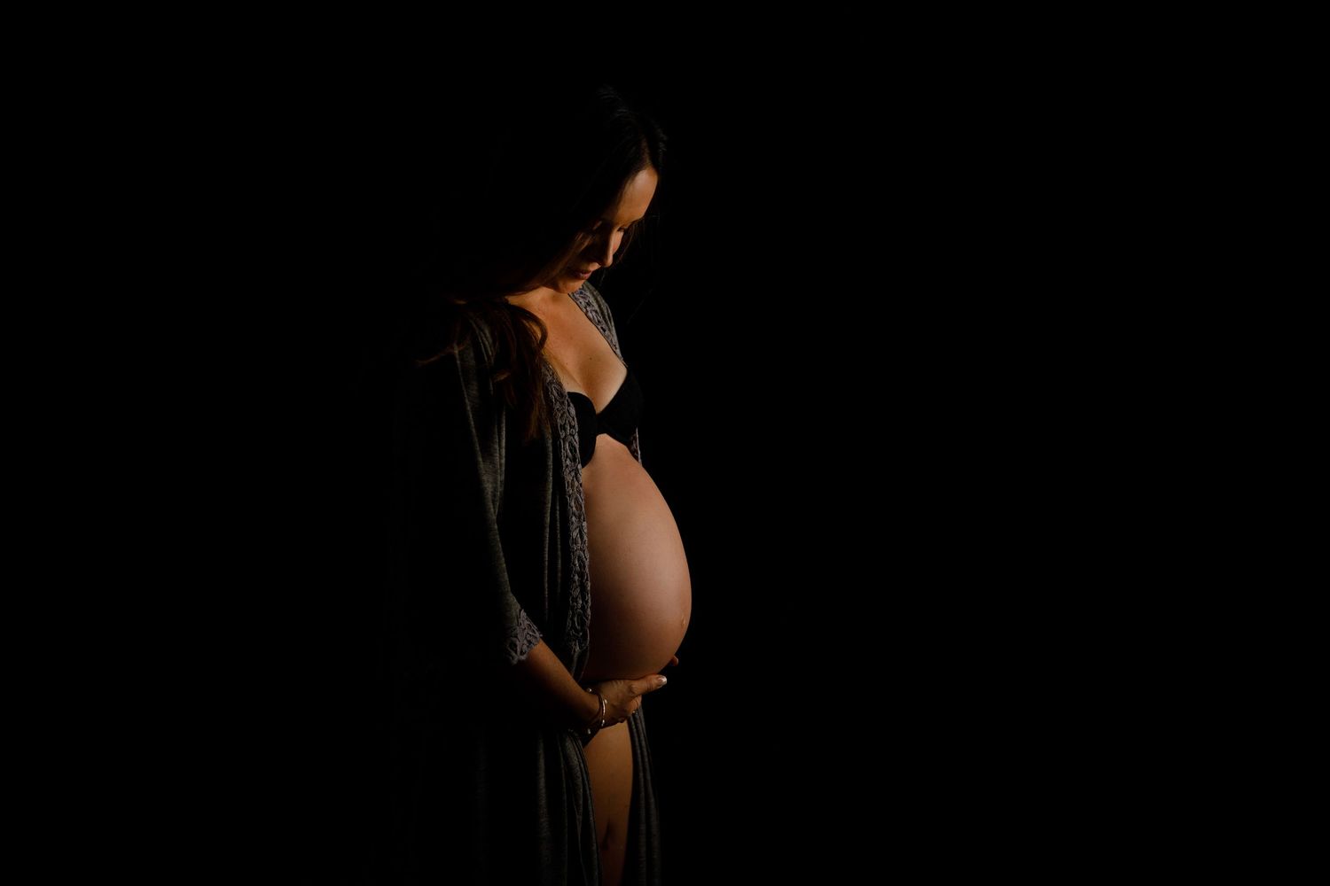 Pregnant woman in dark studio, cradling baby bump in lingerie and sheer robe, dramatic black background maternity photo.