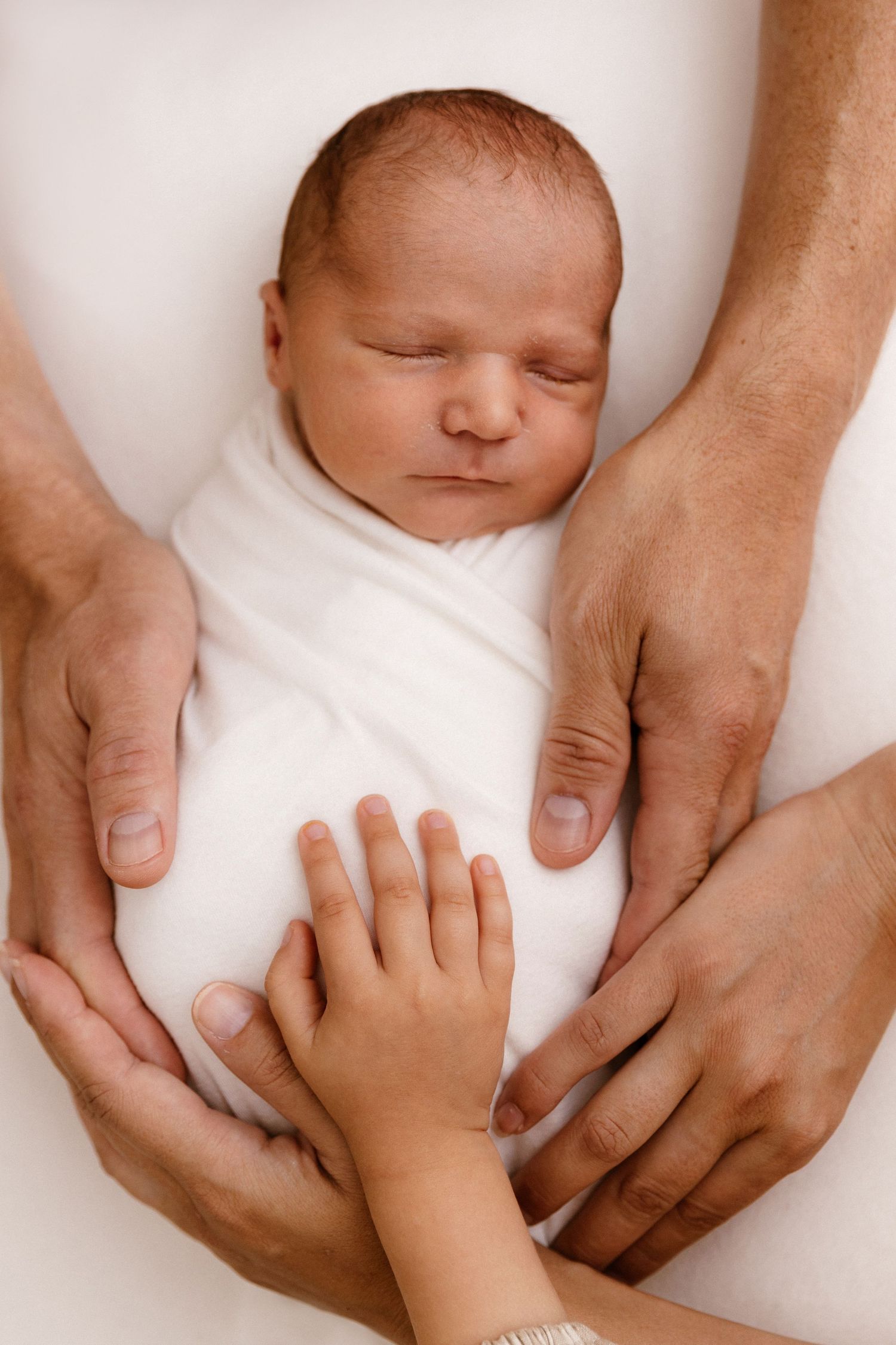 Newborn baby sleeping wrapped in white cloth, surrounded by parents and sibling hands in a tender family moment.
