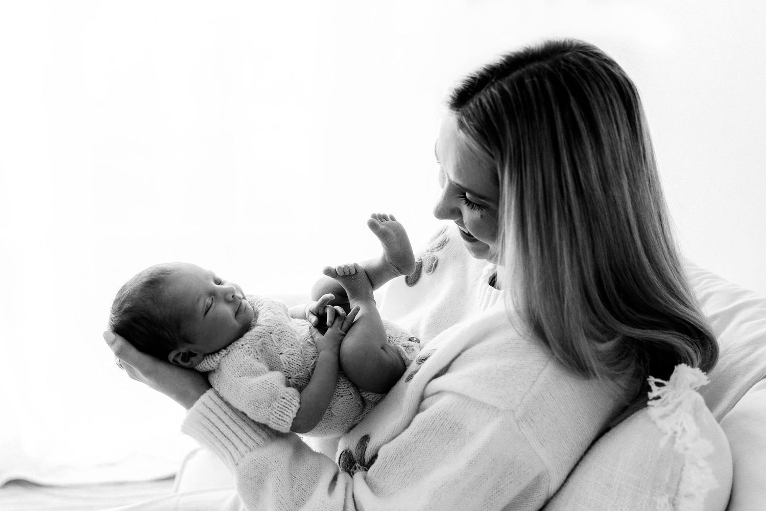 A black and white photo of a mother in a white robe smiling at her newborn baby stretching tiny legs.