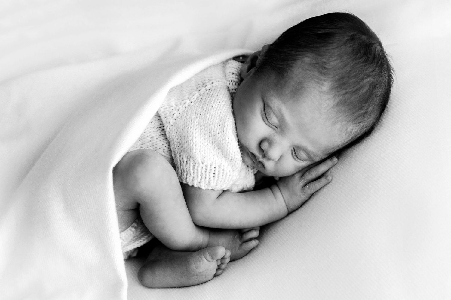 Black and white photo of a newborn baby peacefully sleeping curled up in a soft white wrap.