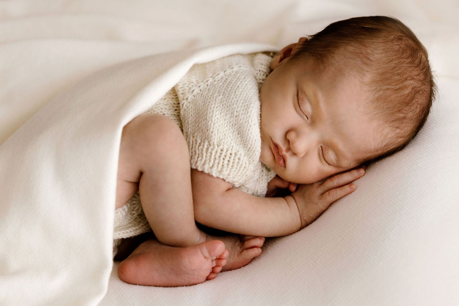 Adorable newborn baby sleeping curled up under a soft white blanket in serene pose.